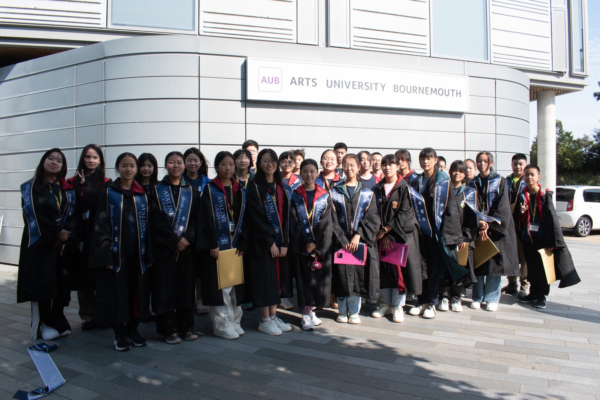 Large group of graduates in black gowns and mortarboards standing outside Arts University Bournemouth building entrance.