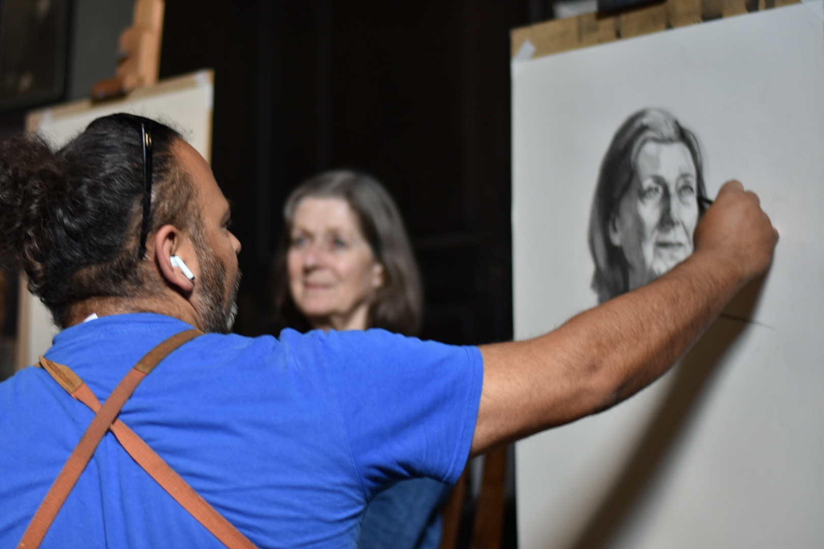 An artist viewed from the back in the process of drawing a woman's face. The model is visible in the background.