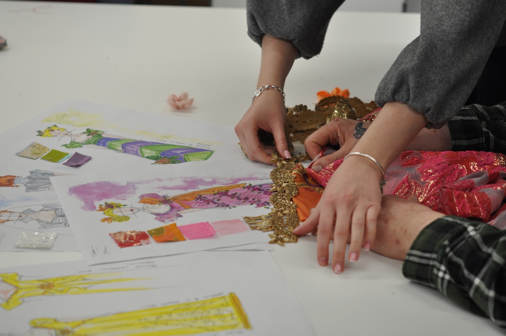 Close-up of two pairs of hands holding gold trim to orange and red fabric samples, while several colourful costume design illustrations lie on the table in front of them.