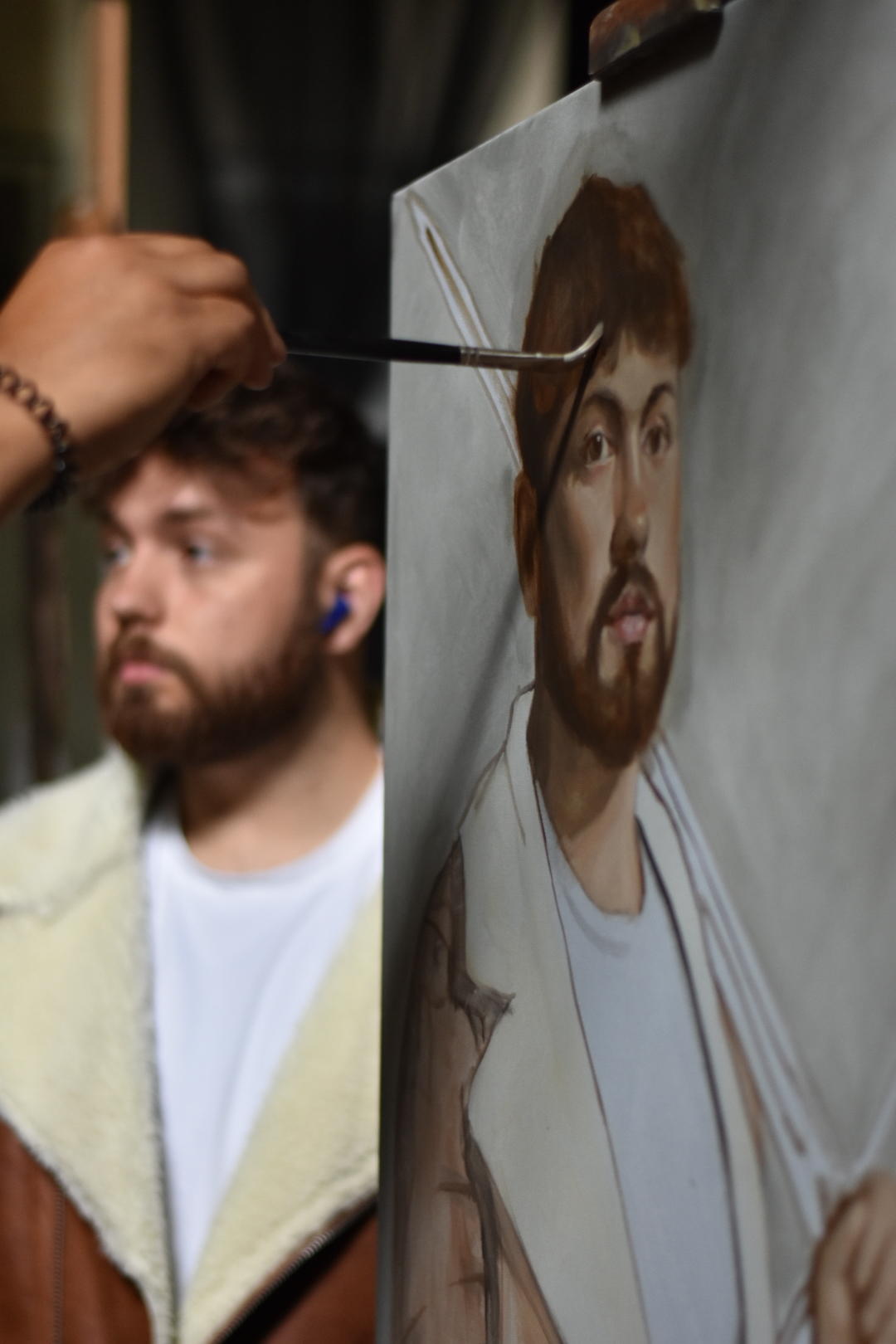Close-up of a portrait of a man with a beard and a fleece jacket being painted. The model is visible on the left.