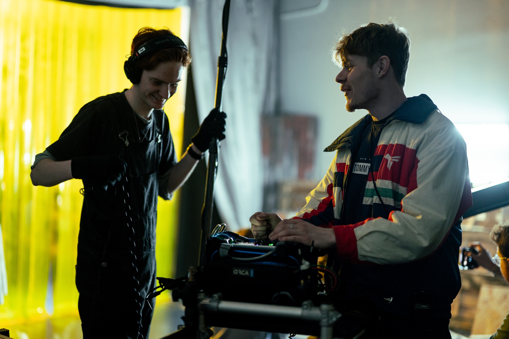 Two men stand beside a film camera on set, with one adjusting equipment while the other holds a boom pole near a yellow backdrop.