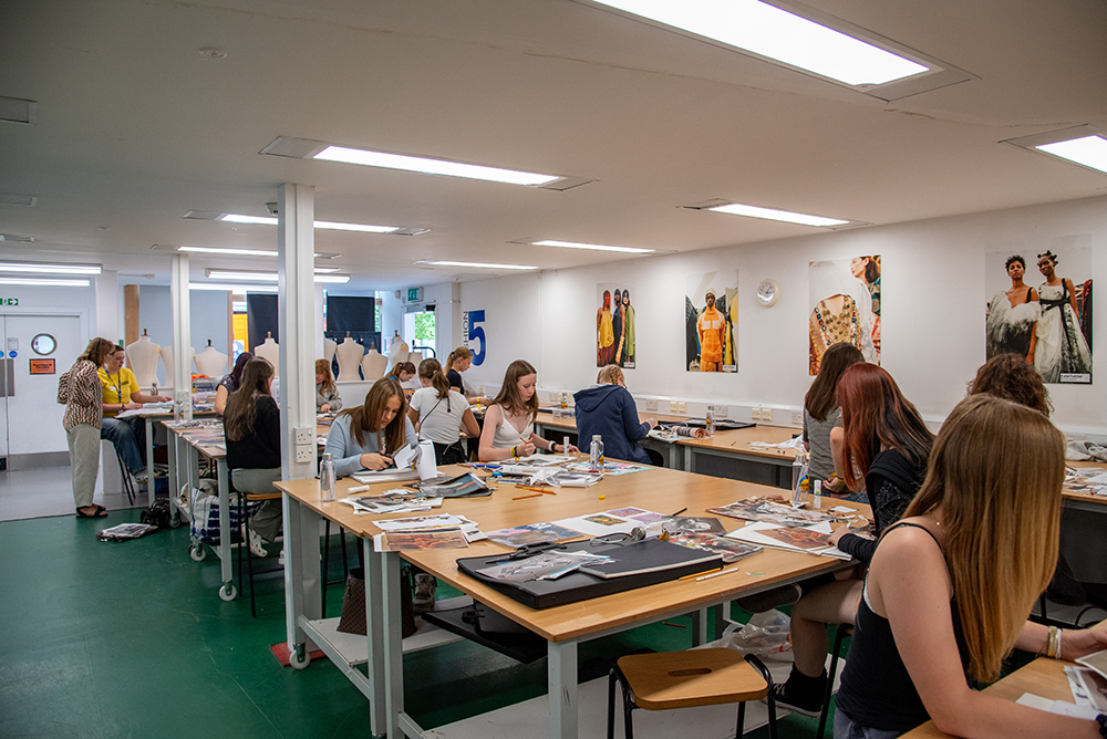 Art classroom with students at wooden tables, green floor, white walls displaying colourful paintings, fluorescent ceiling lights.