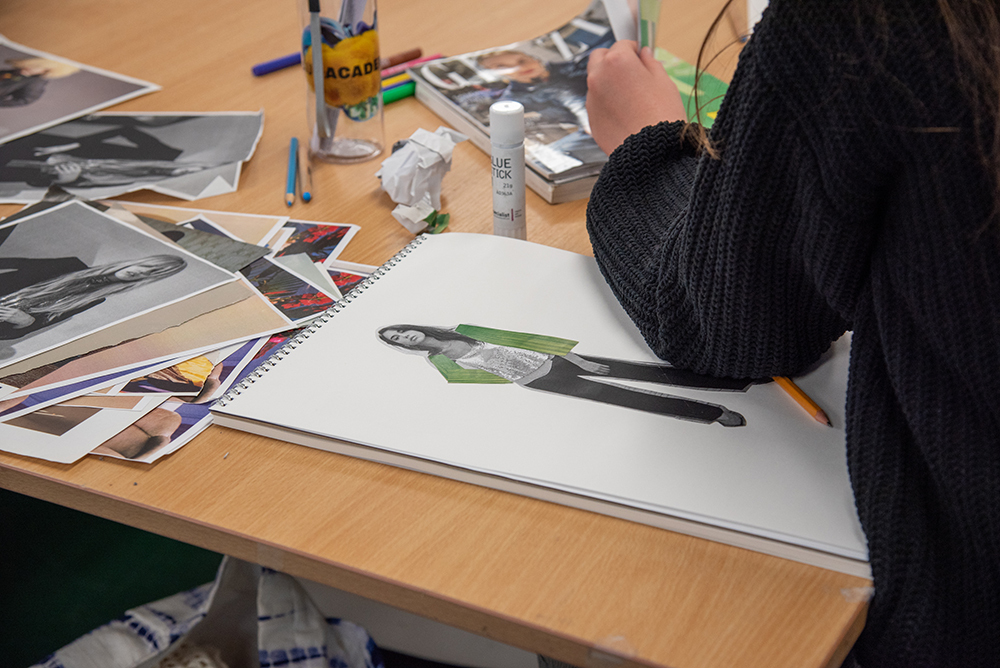 Person in black jumper working at wooden desk with scattered photographs, art supplies, and white sketchbook containing green illustration.