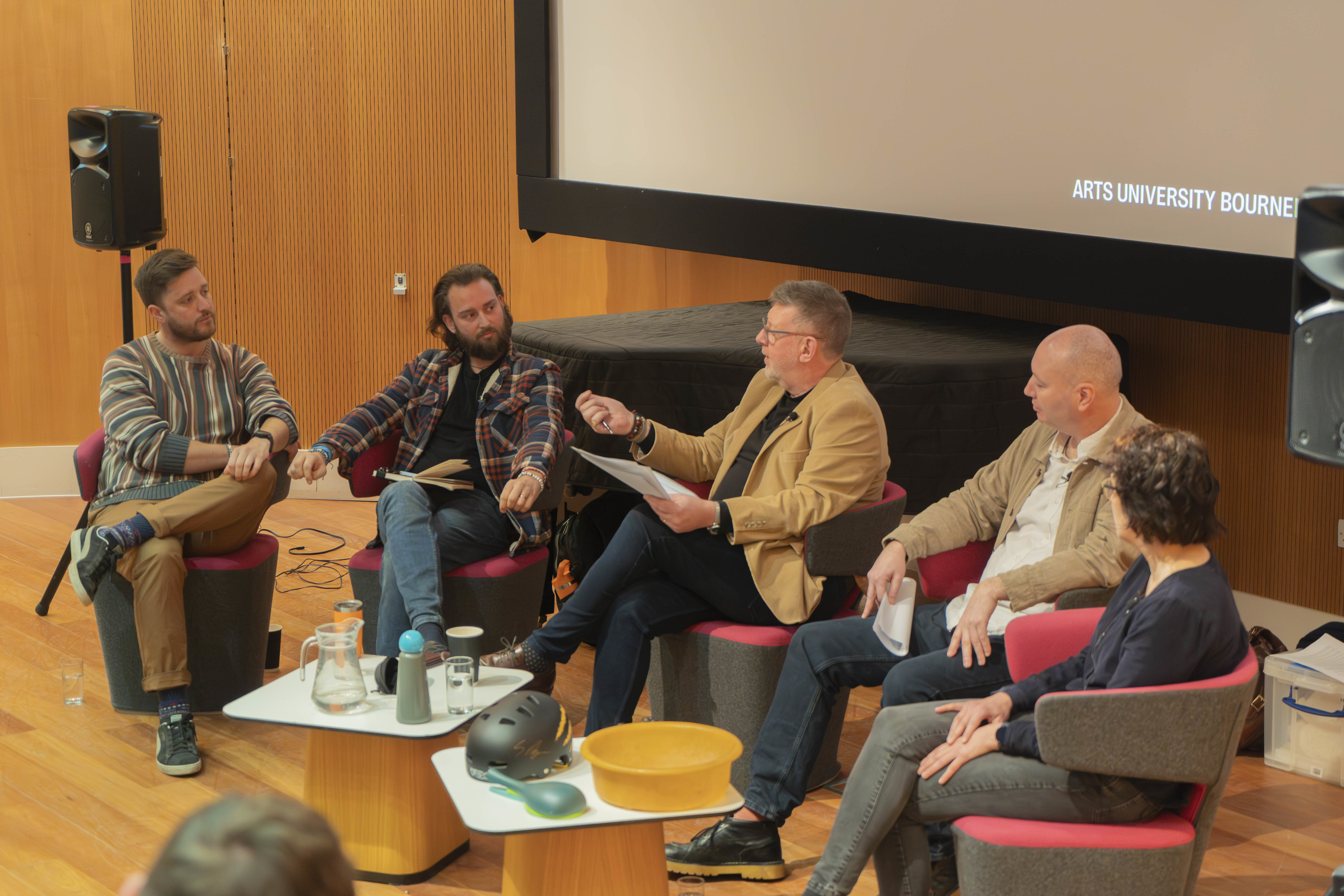 Brothers Make, T Ryan, W Strange and L Dennis sitting in a semicircle in a lecture theatre, conversing during the recording of a live podcast.