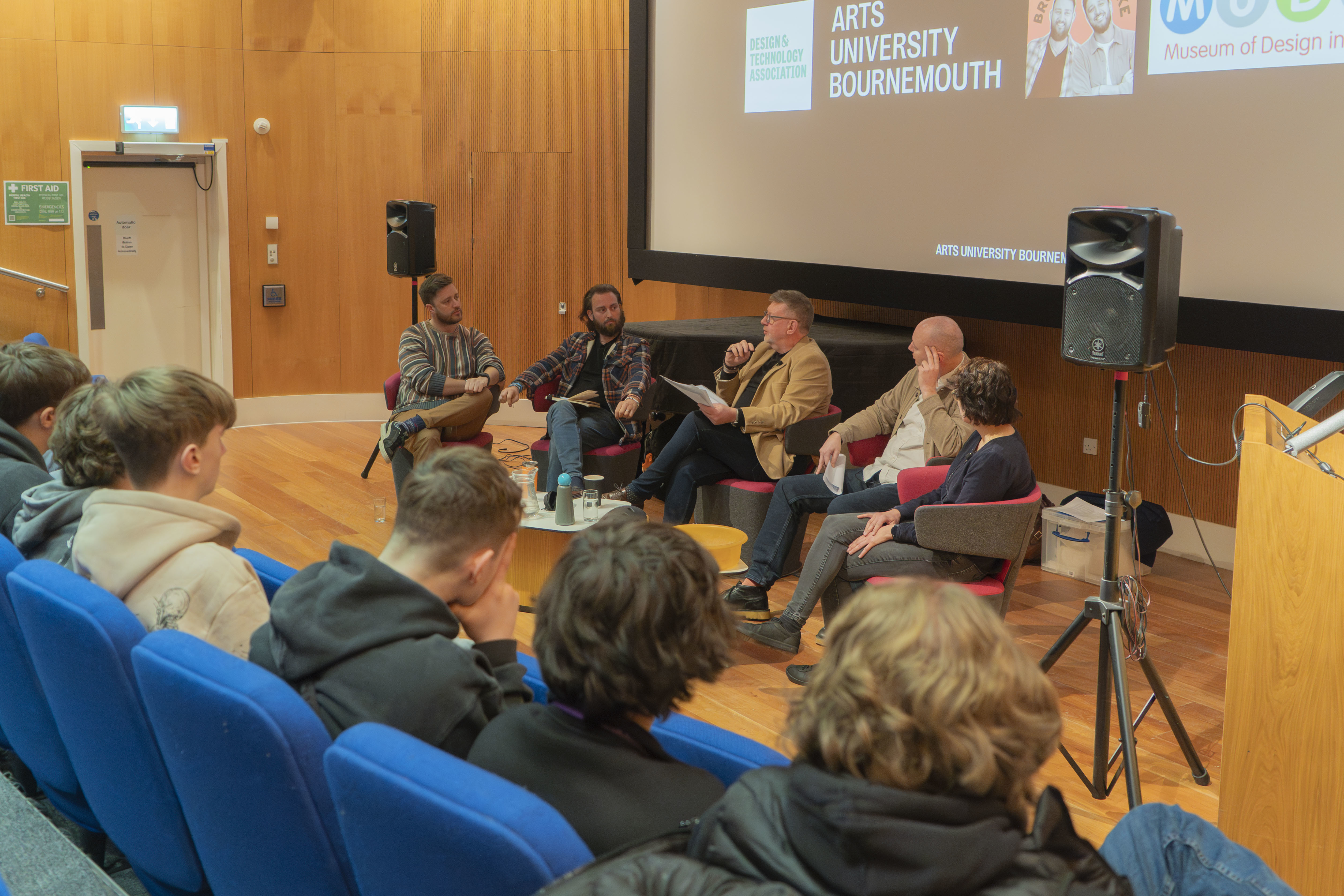 Brothers Make, T Ryan, W Strange and L Dennis sitting at the front of a lecture theatre and presenting a live podcast to a group of young people. Some young people are viewed from the back, sitting in the audience.