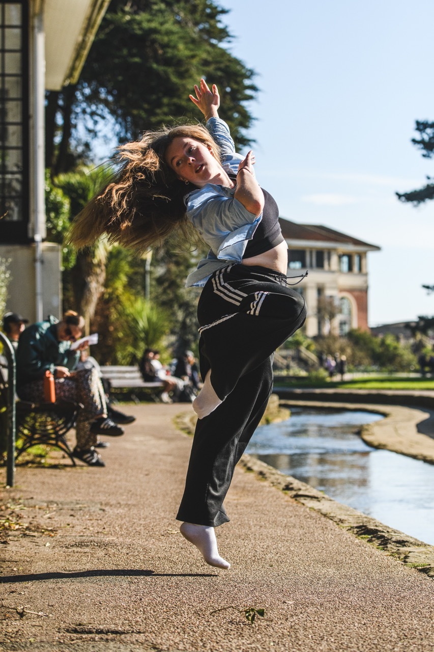 Woman mid-jump with arms raised, wearing grey top and black trousers, on pavement beside water feature with buildings behind.