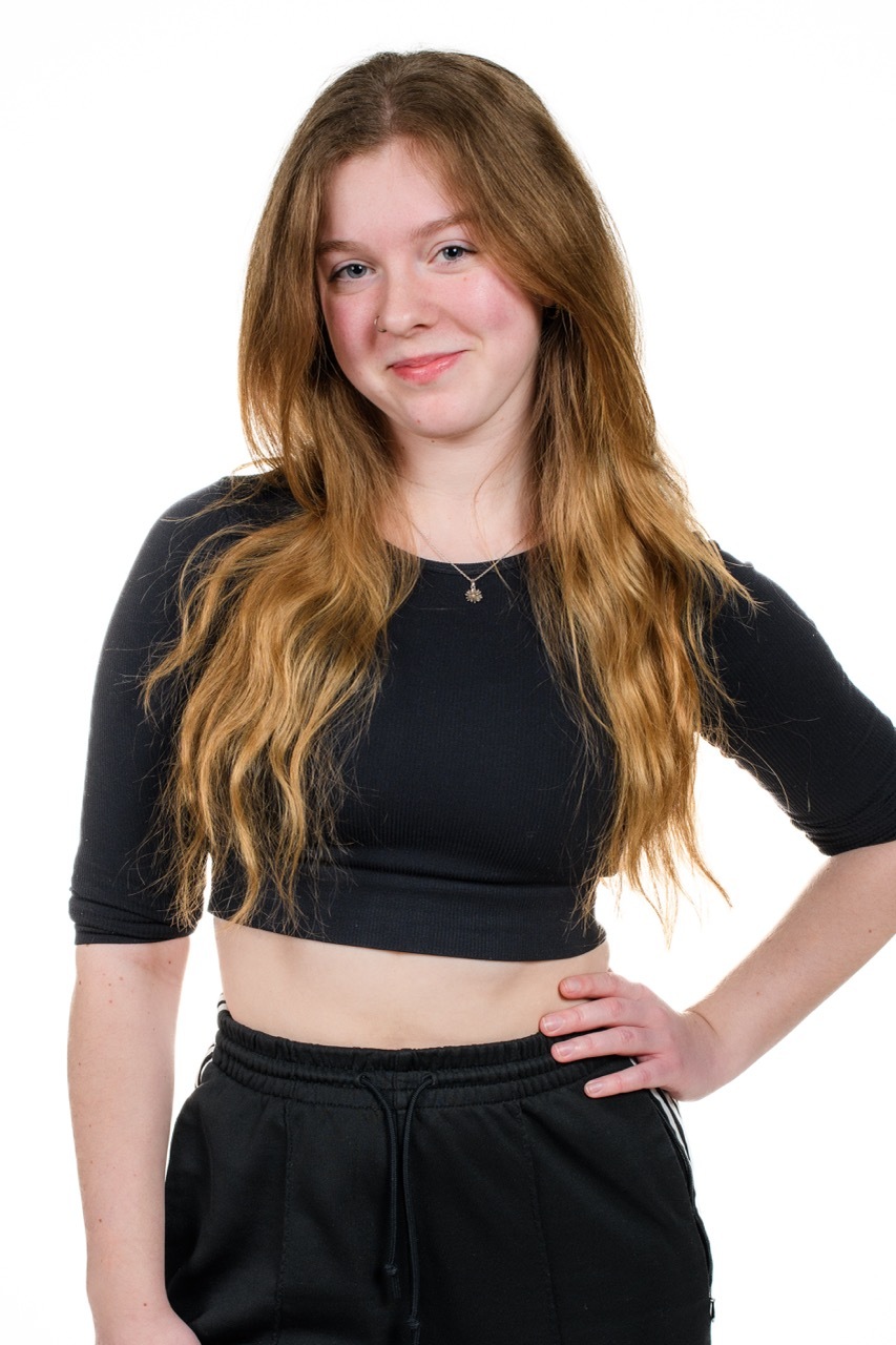 Young woman with long blonde hair wearing black crop top and black joggers, hand on hip, smiling against white background.