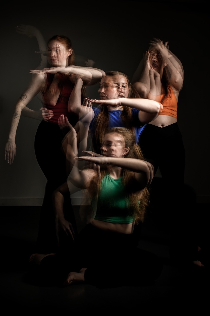 Four dancers in colourful tops creating interlocking arm formations against dark background with dramatic lighting.