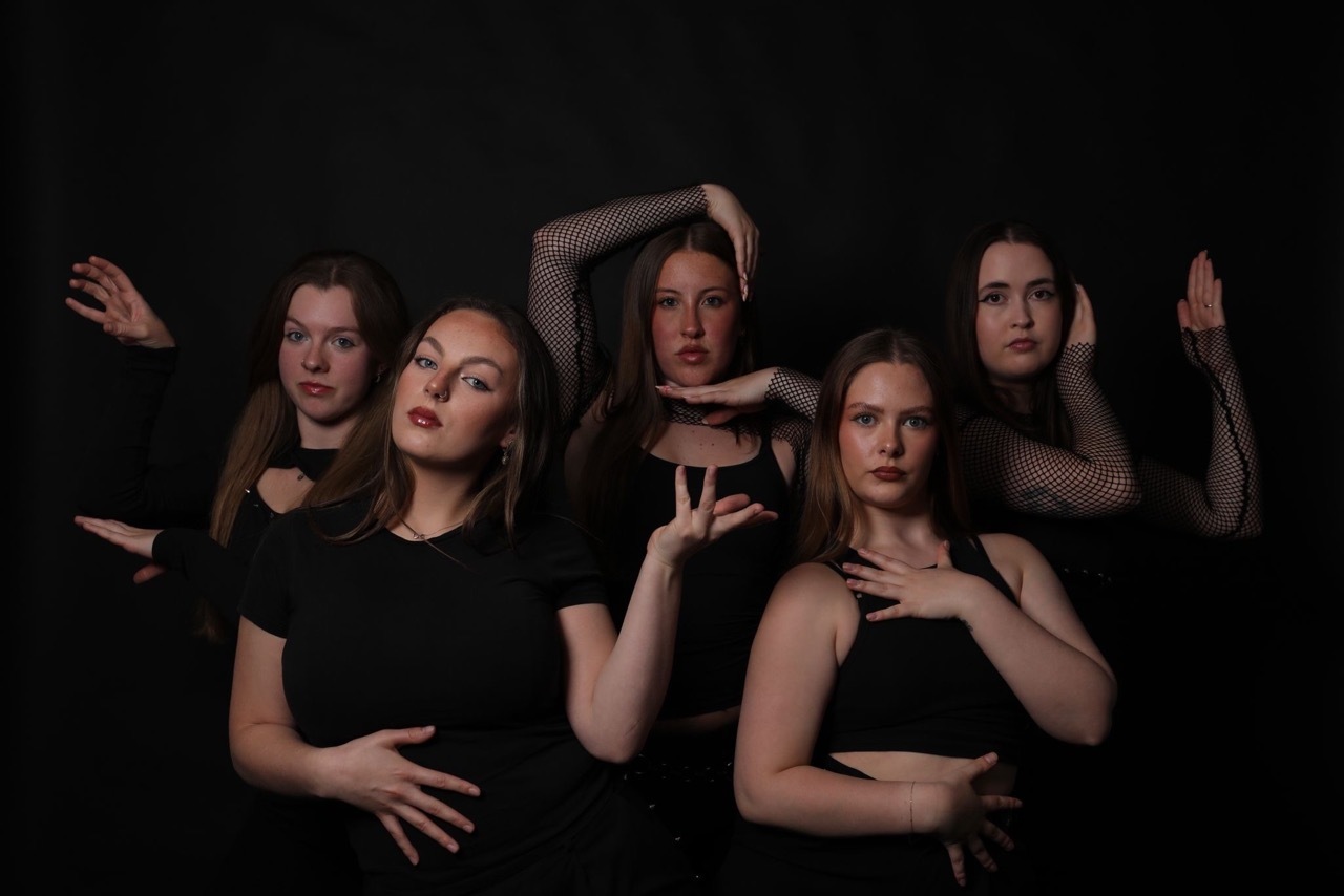 Five women in black clothing posed with raised hands and expressive gestures against dark background.