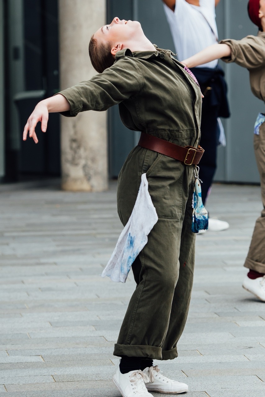 Person in olive green jumpsuit and brown belt leaning backwards dramatically on street, head tilted up, one arm extended.