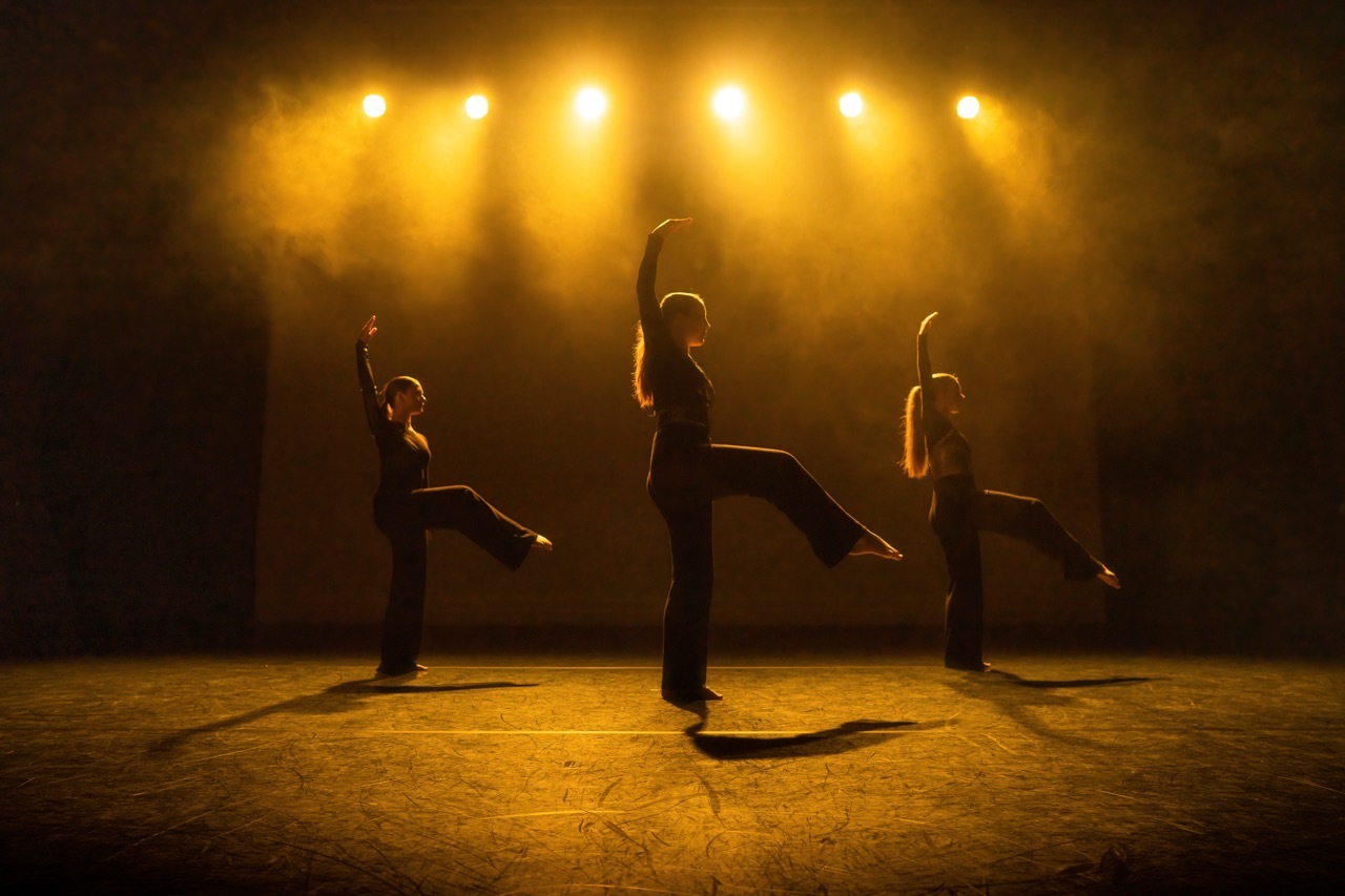 Three dancers in silhouette against bright yellow stage lights, arms extended, performing on wooden floor with dramatic backlighting.