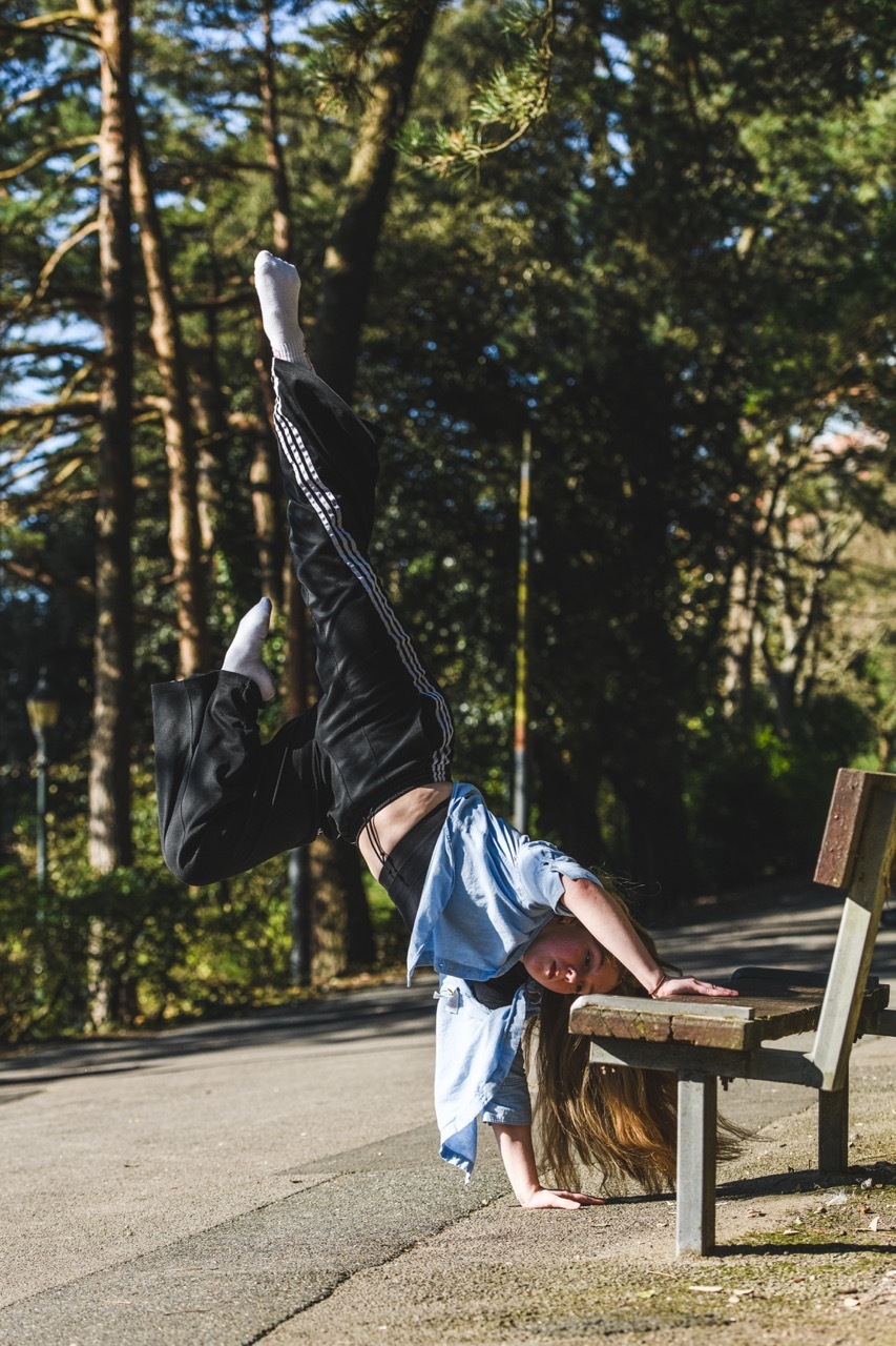 Person doing handstand with legs over wooden bench in park, wearing black jacket and blue jeans, surrounded by green trees.