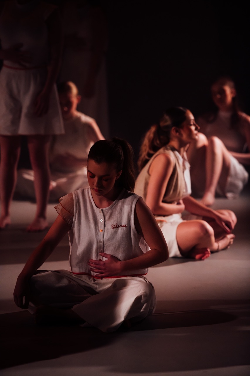 Group of people sitting on floor in dimly lit room with warm orange lighting, wearing casual white clothing.