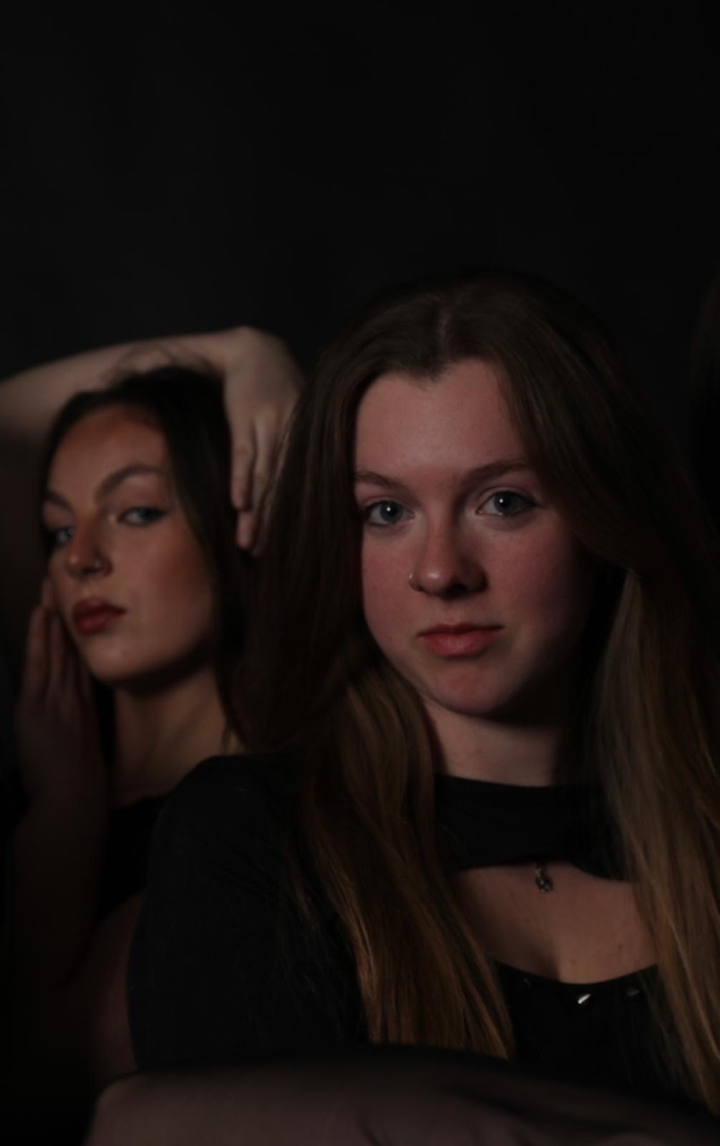Two young women with long hair in dim lighting, one with hand raised to her head, both wearing dark clothing against black background.
