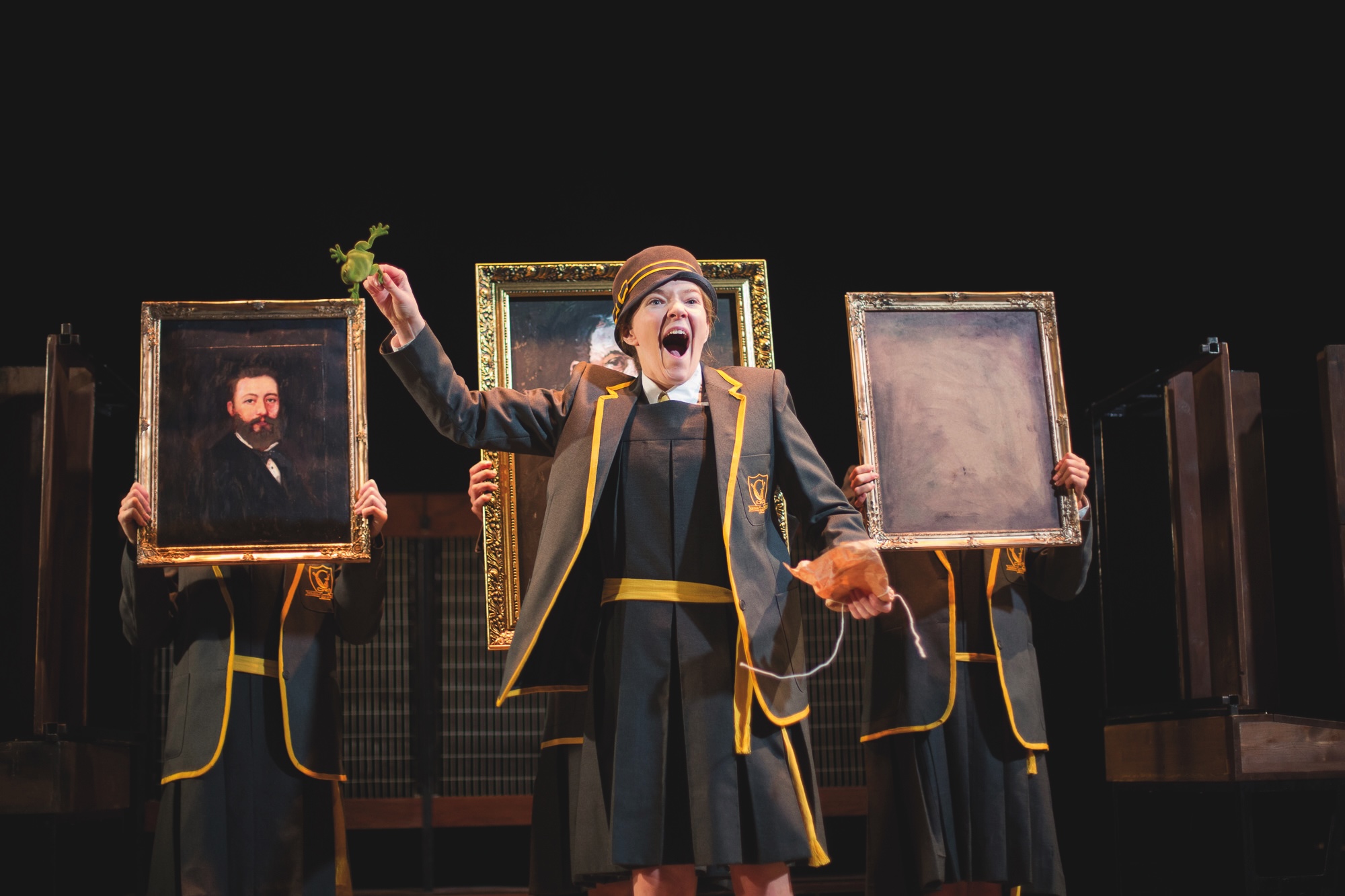 A performer in a dark costume with gold trim stands centre stage holding up a small sprig, flanked by two people holding framed portrait paintings.