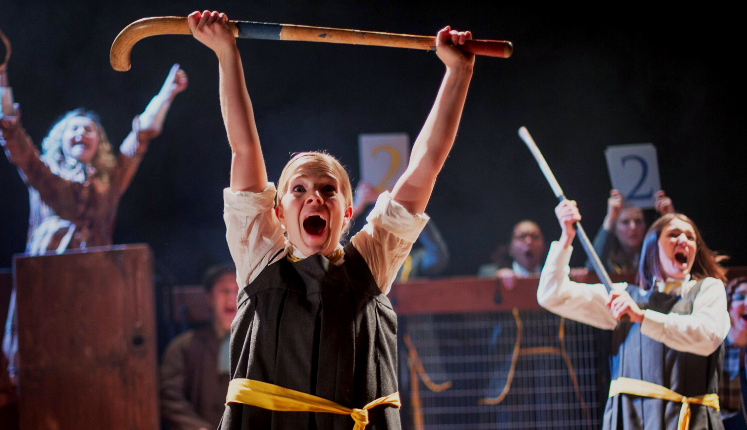 Young person in white shirt and dark waistcoat holds curved wooden stick above head with both hands. Other performers with sticks visible in background on stage.