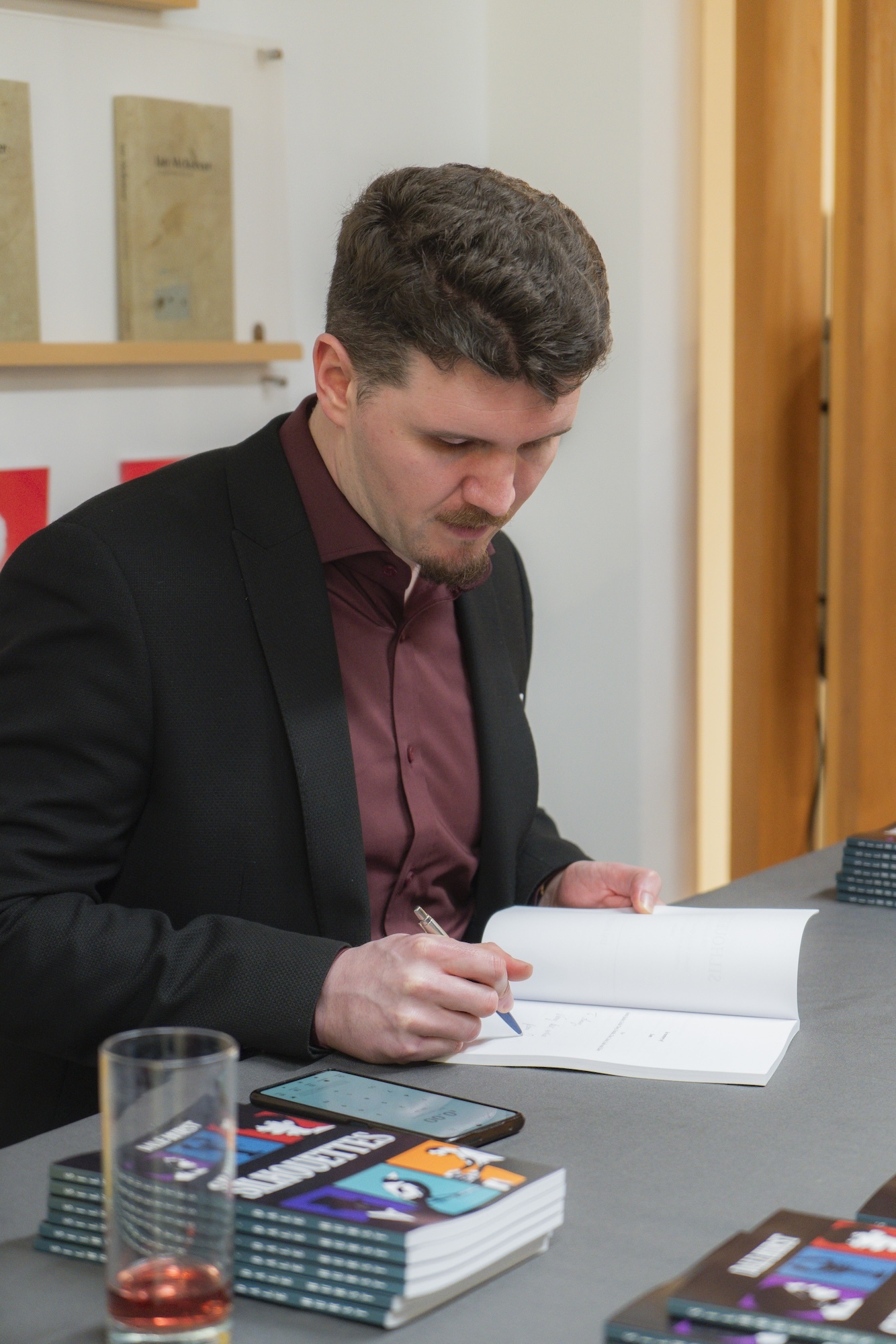 D Hurst signing book at table with stack of books and glass nearby at TheGallery, Arts University Bournemouth