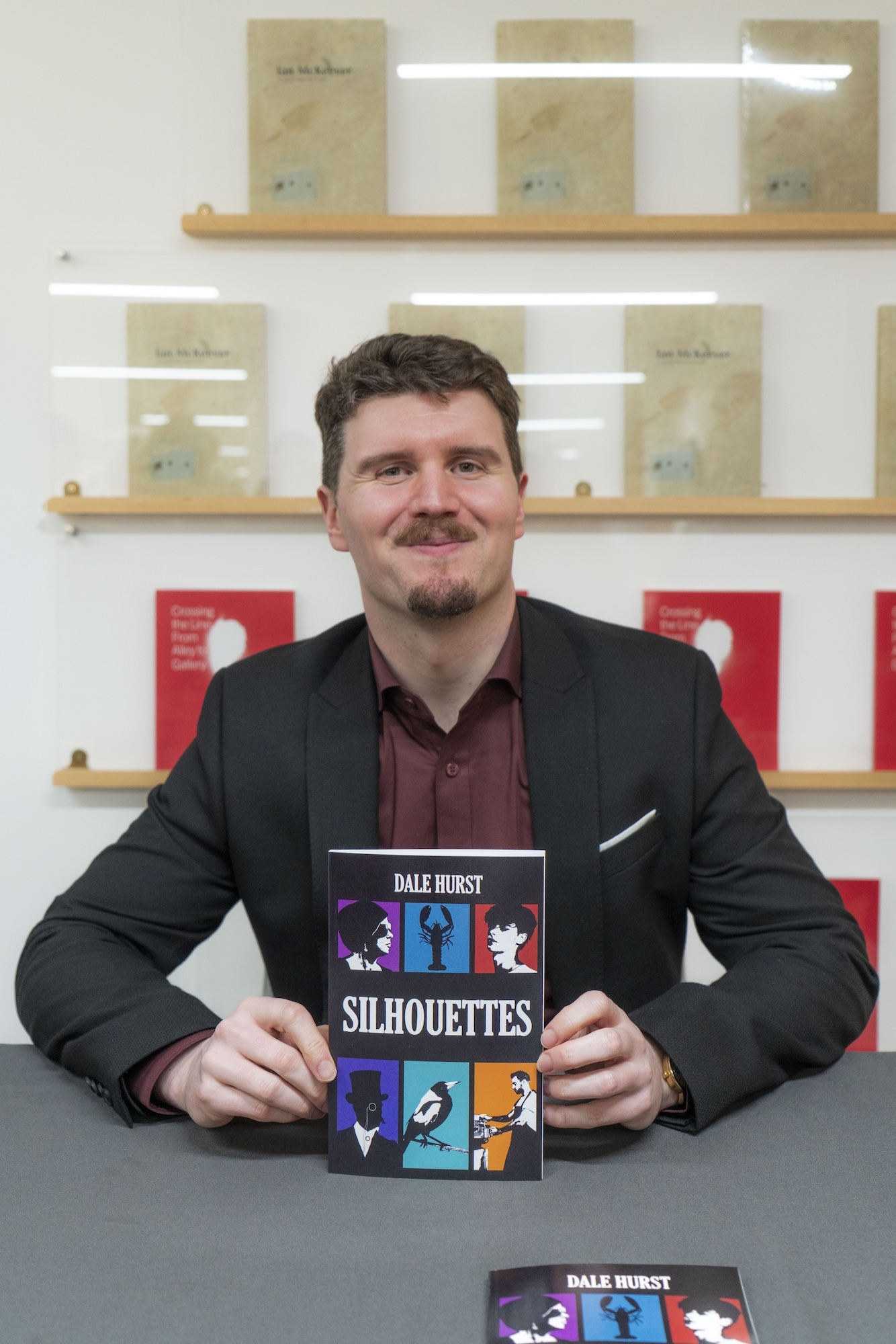 D Hurst holds "Silhouettes" book at signing table with bookshelf behind at TheGallery, Arts University Bournemouth