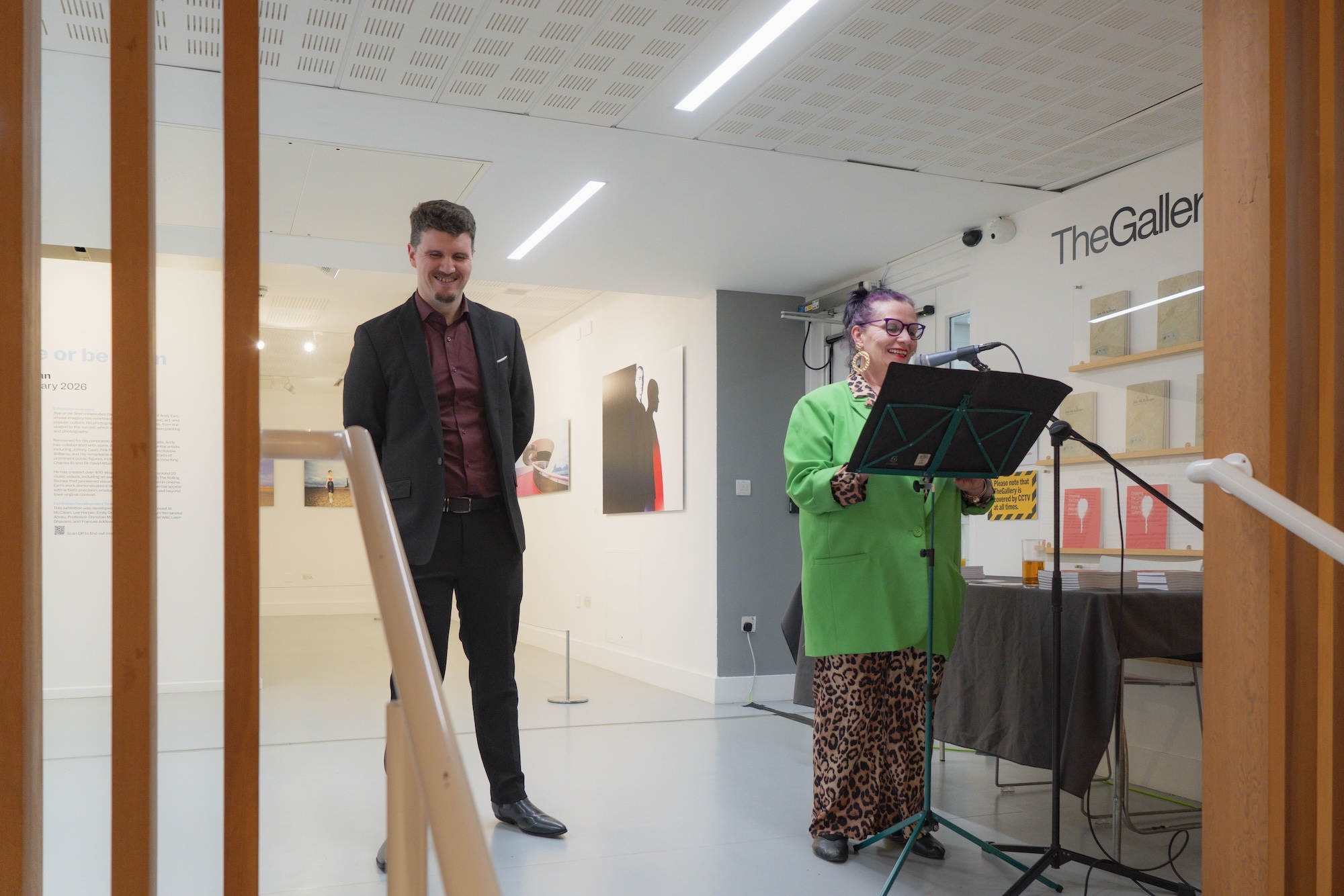 Two people in modern gallery space with white walls and orange columns. Man in dark suit stands left, woman in bright green jacket reads from lectern right.