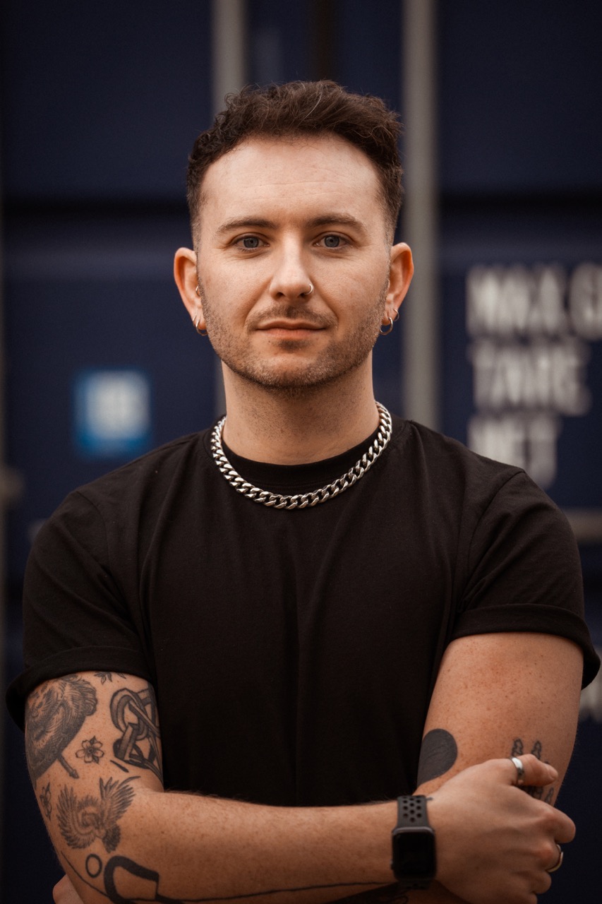 Portrait of Dan Branfitt with short dark hair, wearing a silver necklace and dark t-shirt.
