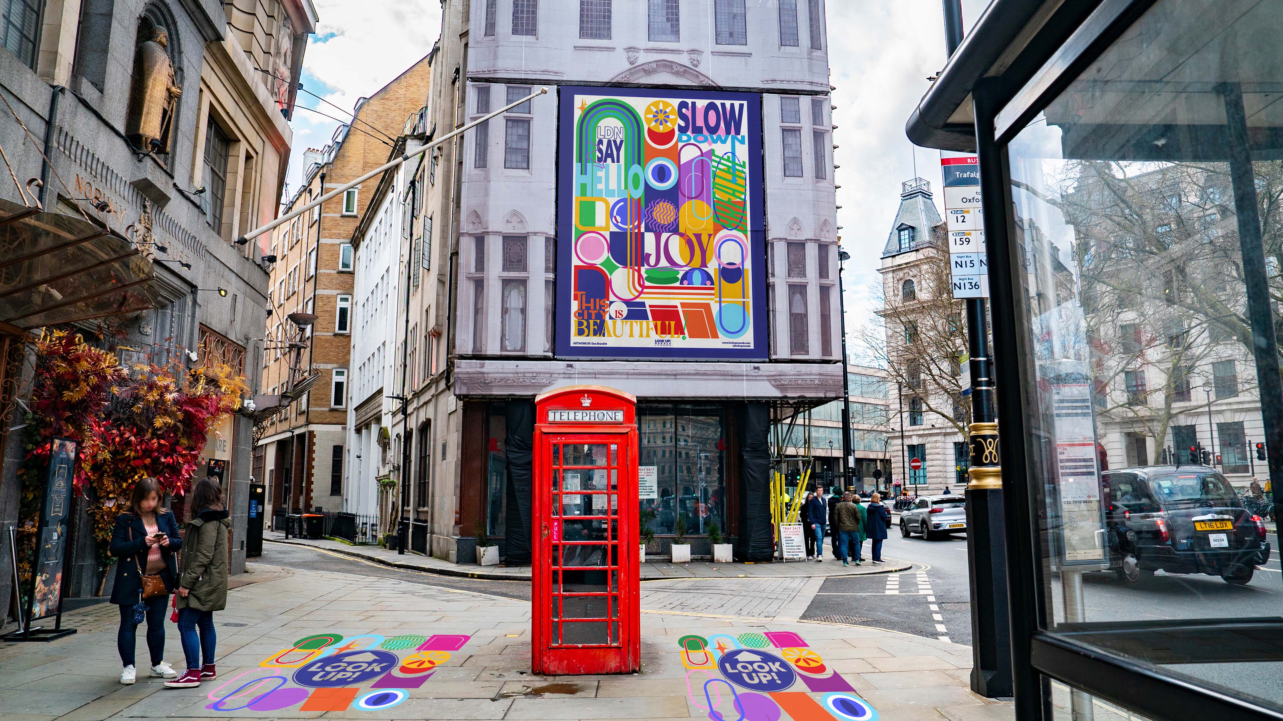 Abstract, colourful graphic design shapes on the floor and billboard of a building in a street, red telephone box in the middle.