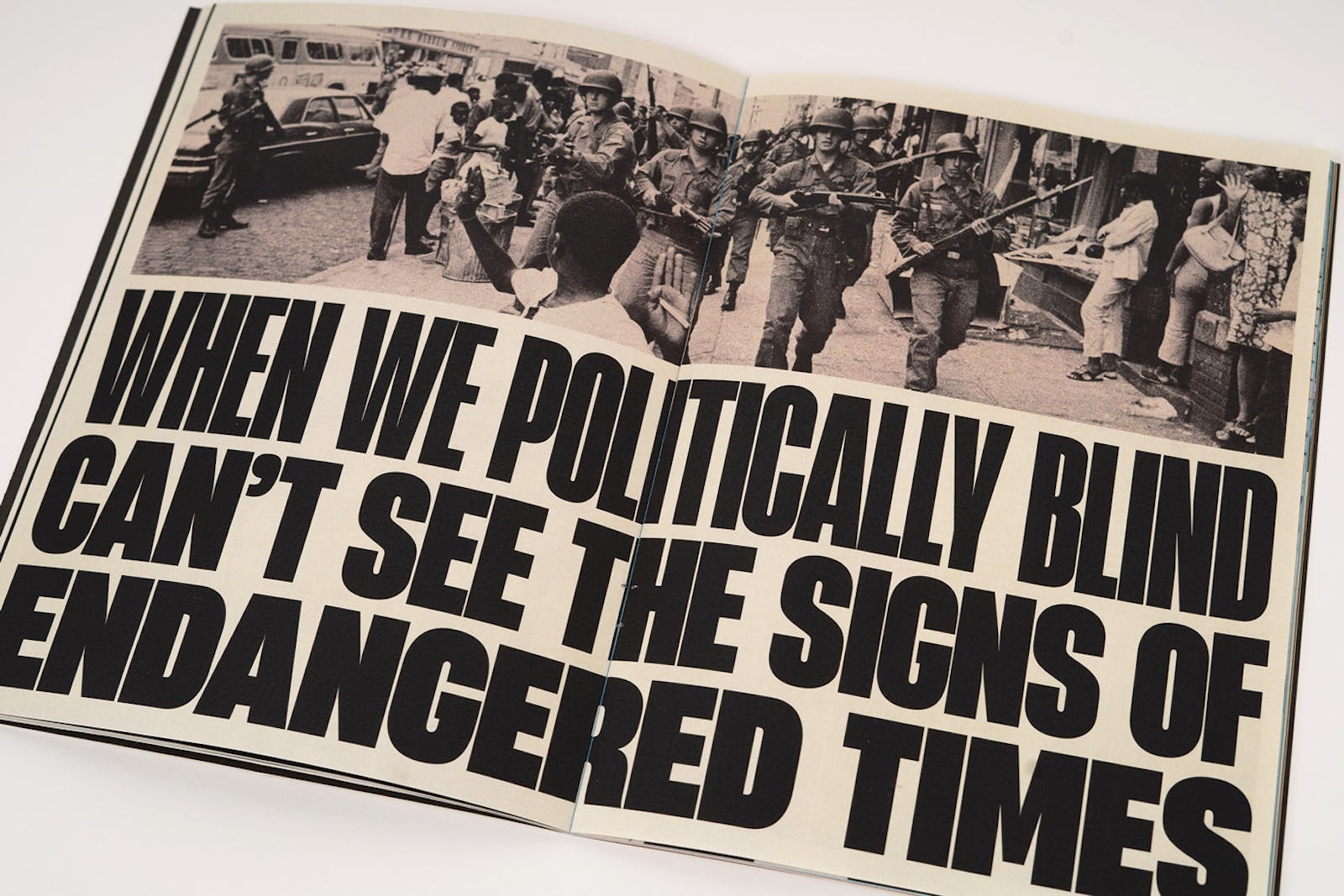 Book open to a double page spread showing a black and white photograph of armed soldiers marching down a street. Bold headline text appears underneath, reading "When we politically blind can't see the signs of endangered times".