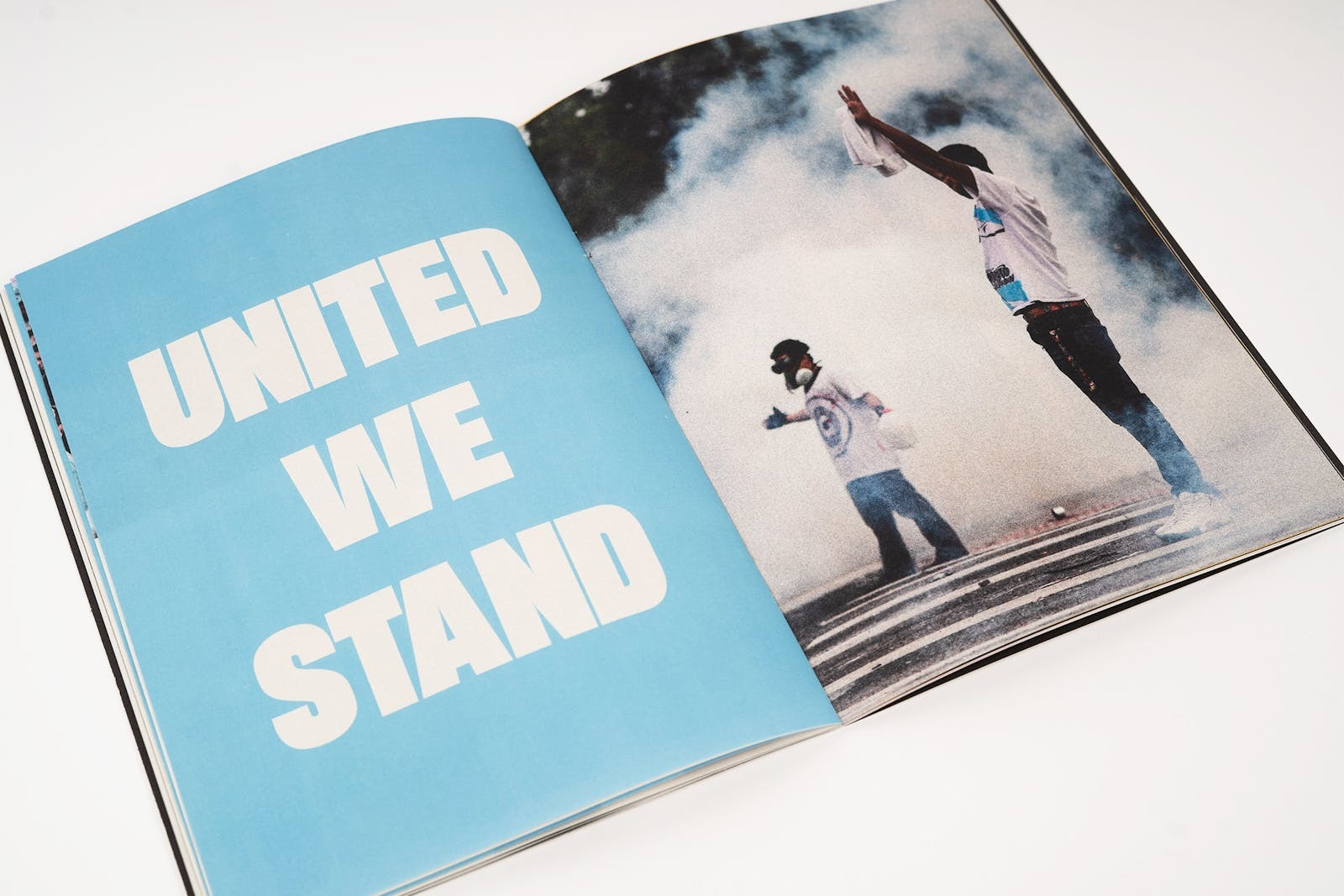 Excerpt from a physical magazine, the headline reads 'United we stand' the photo overleaf depicts two people of colour surrounded by smoke and holding their hands in their air, one person has a white piece of textiles in their hand