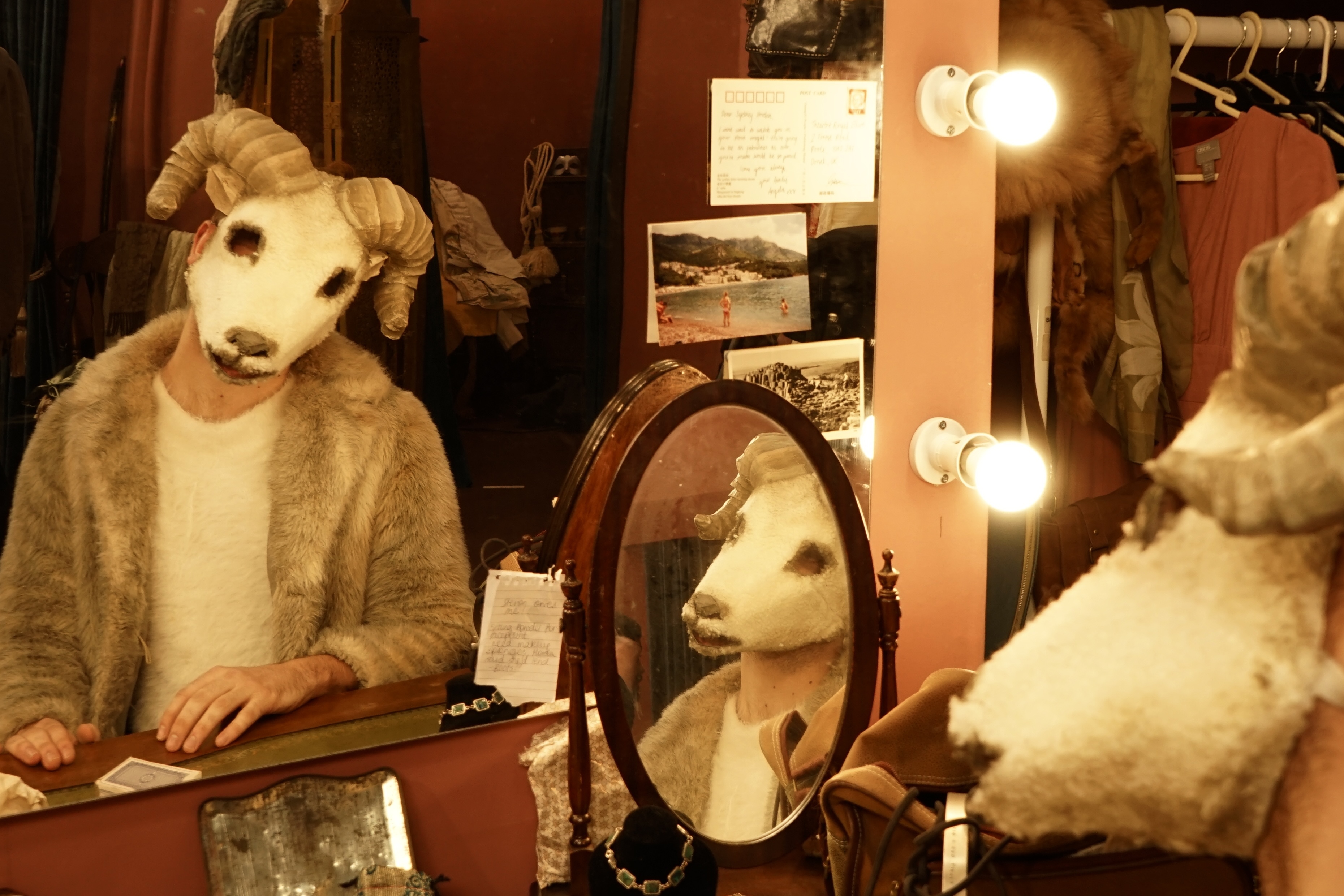 A BA (Hons) Acting student wearing a fur-style coat and a mask of a sheep's head, viewed in a large dressing room mirror with flashbulbs around it.