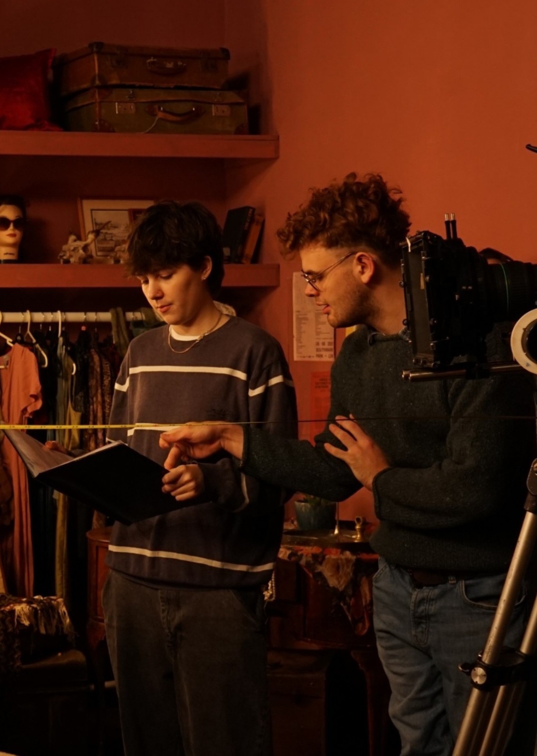 Two BA (Hons) Acting students conferring over a script on a film set in a dressing room, with a rail of clothes visible in the background.