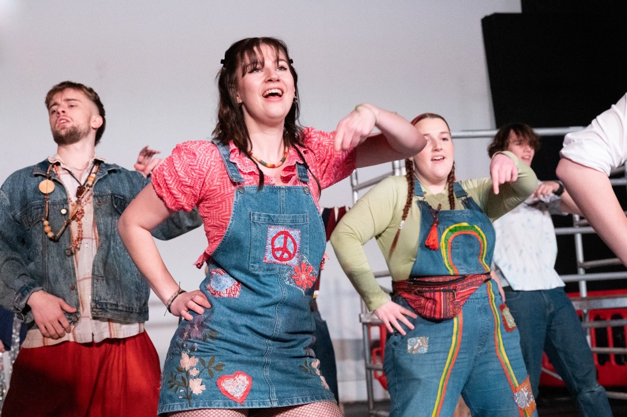 Woman in denim dungarees with peace symbol and patches performs on stage with costumed actors in theatrical production.