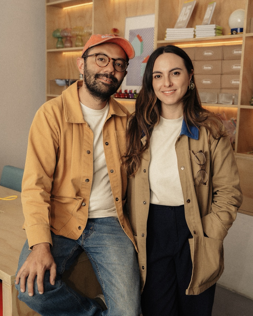 A man and a woman wearing casual jackets stand together in a room with wooden shelves and storage drawers behind them.