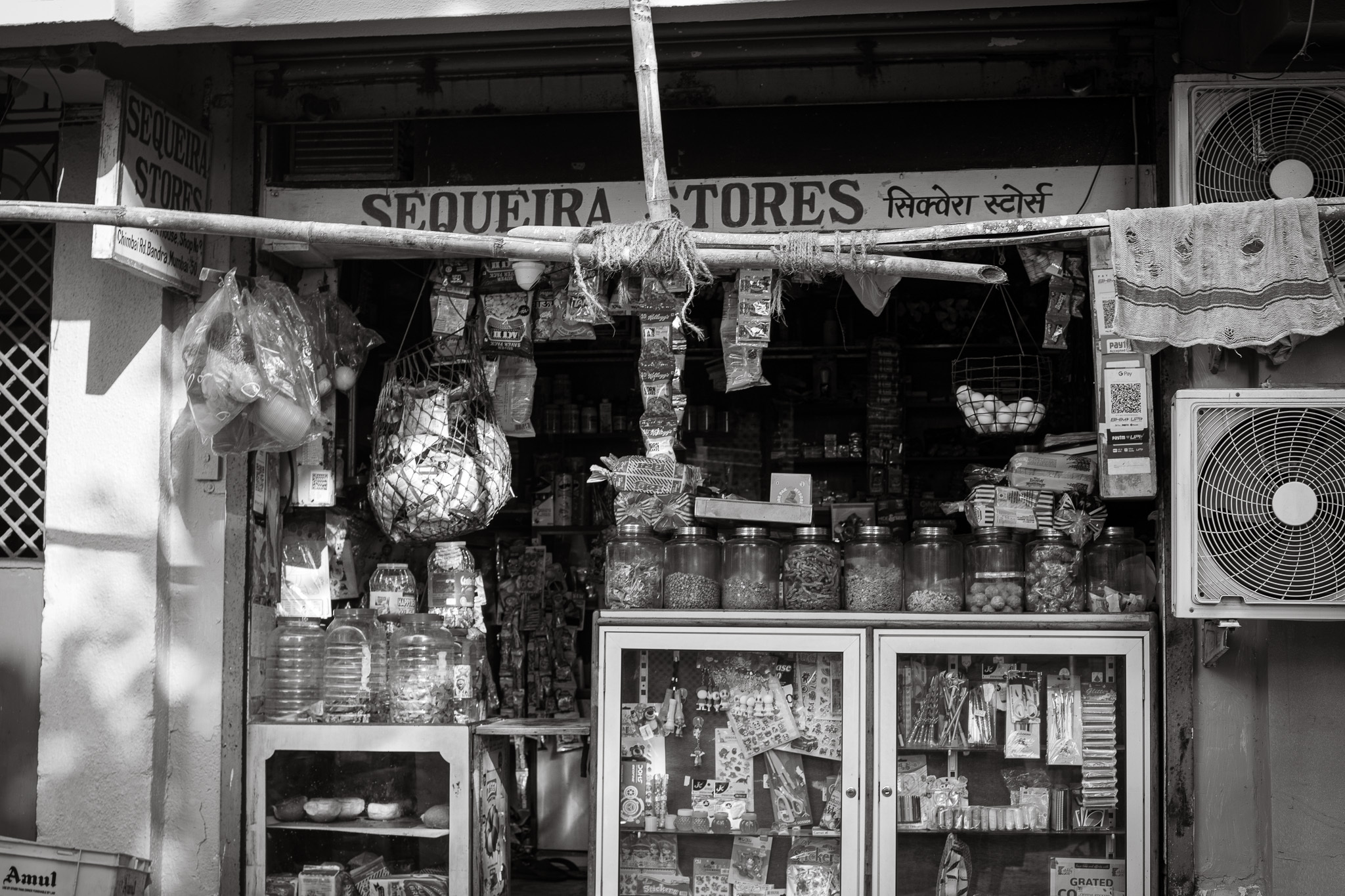 Black and white image of small shopfront with "SEQUEIRA STORES" sign, displaying various goods on shelves and hanging items.