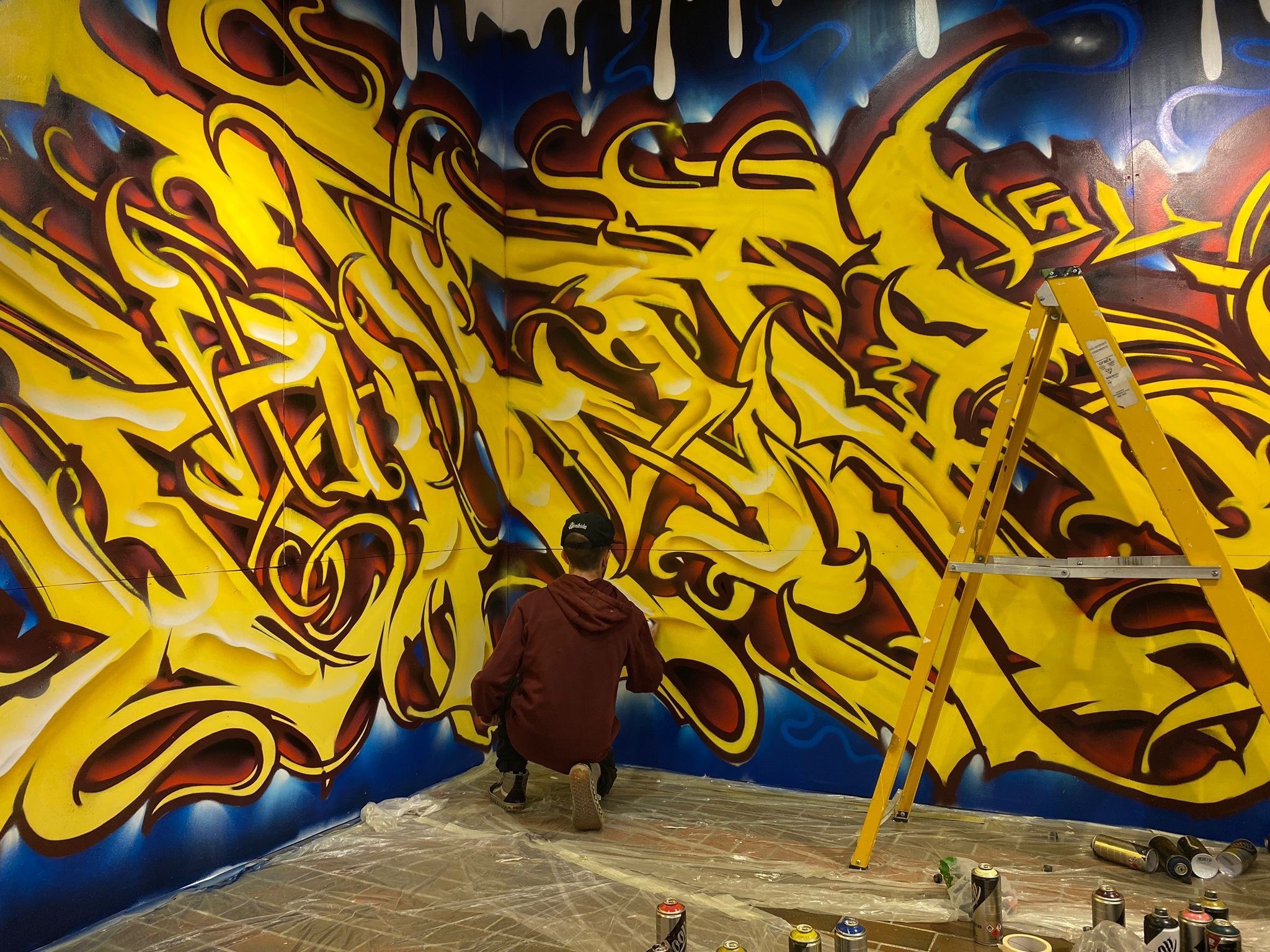 A street artist kneeling down in the corner of a gallery wall, which is decorated with street art.