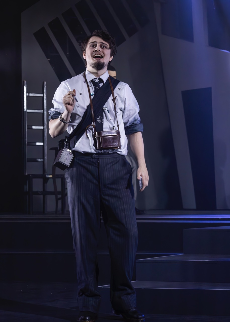 Male performer on stage in white shirt, dark trousers, and leather satchel, holding conductor's baton with ladder visible in background.