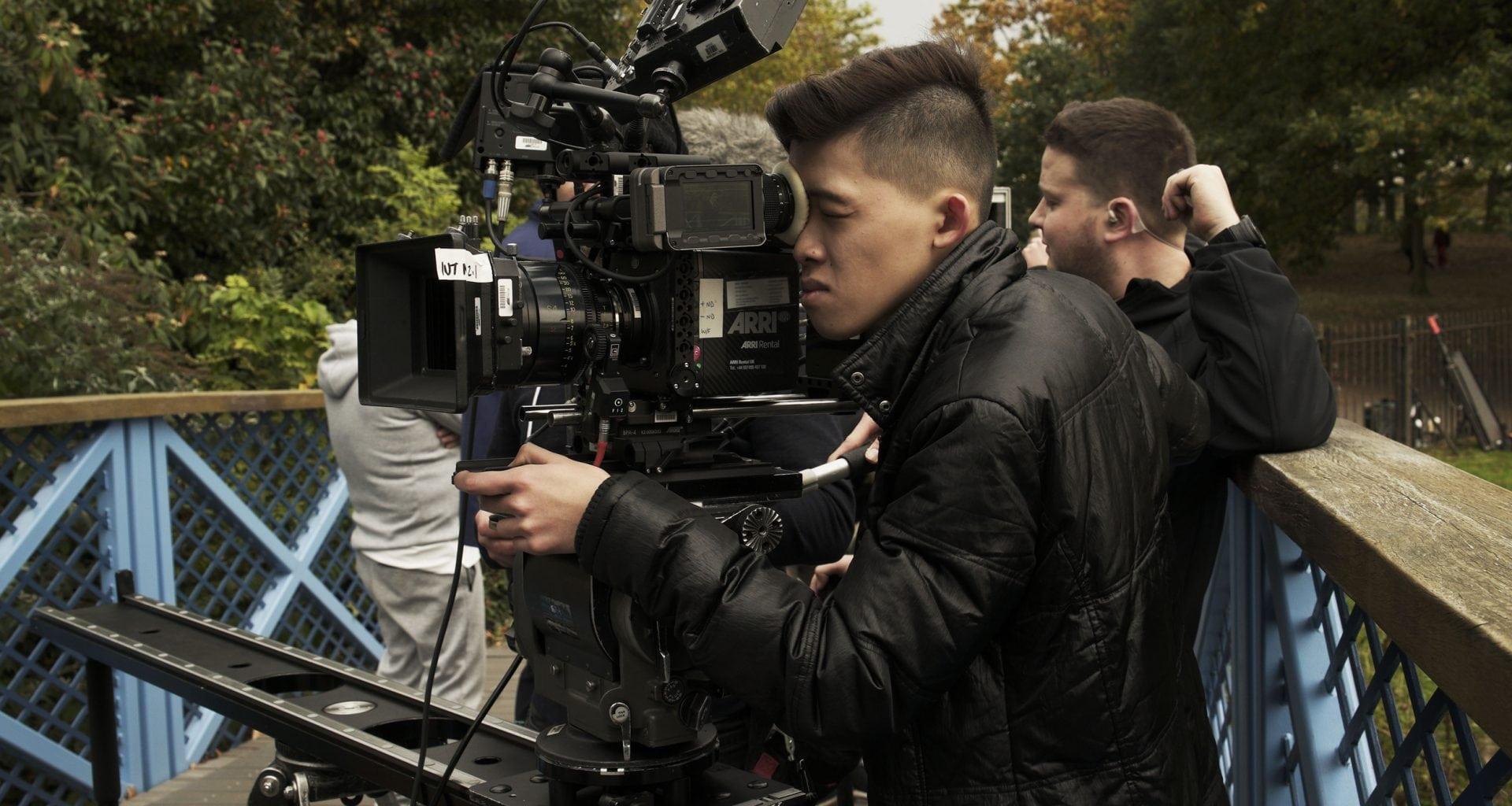 Darius Shu stands on a bridge with a film crew, peers through and holds a camera, filming on the set of Secret Child
