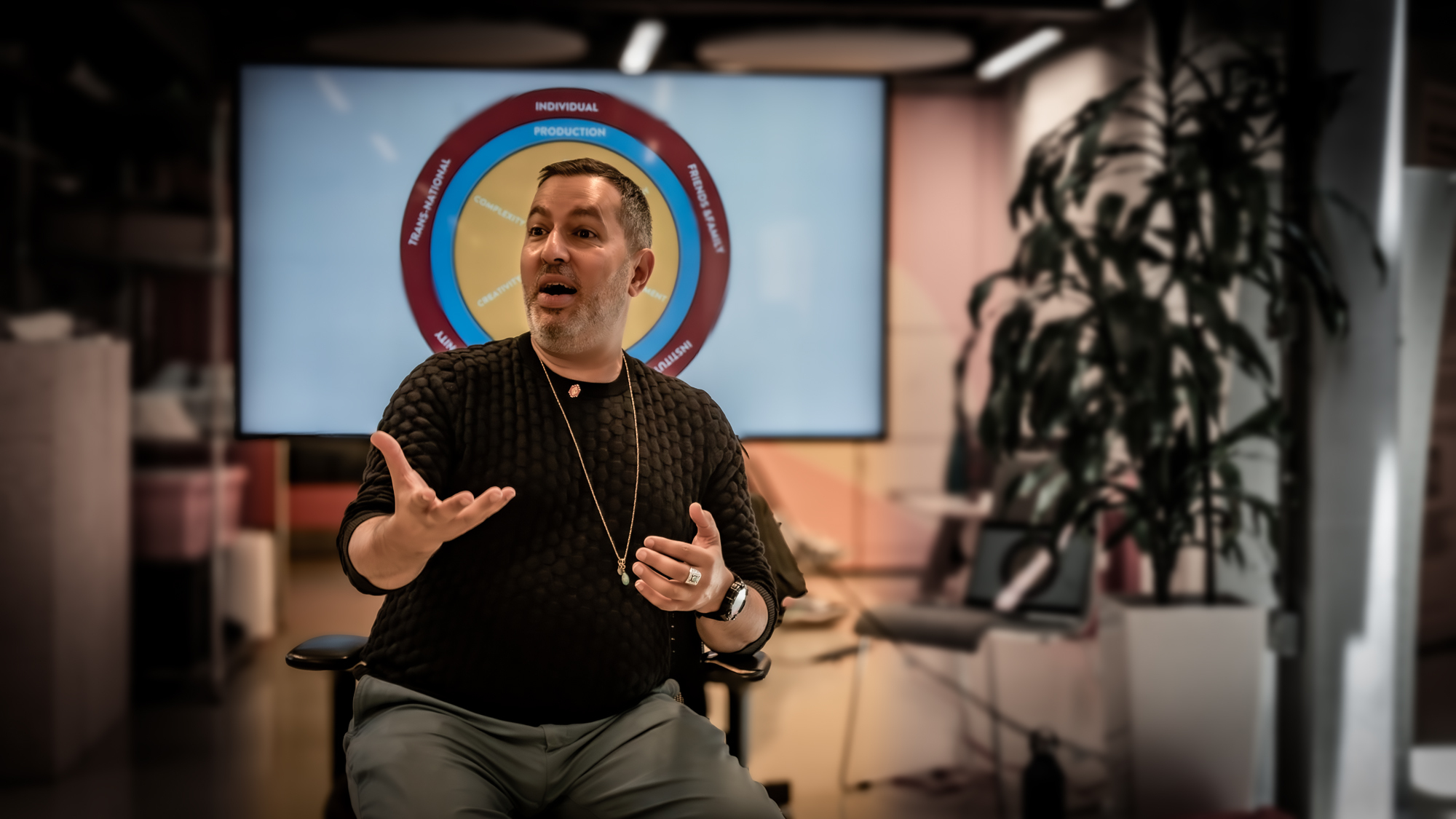 Man in dark jumper gesturing with hands whilst seated in office space with screen displaying circular logo behind him and plant visible.