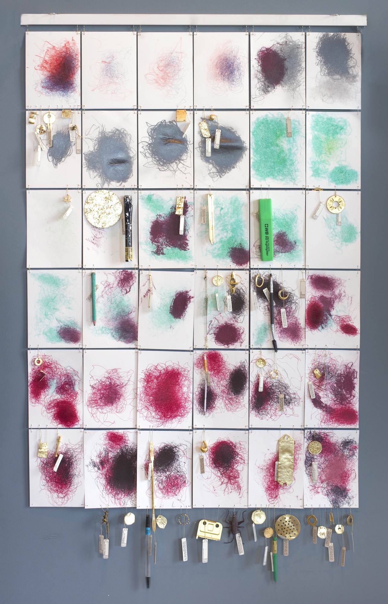 A wall-mounted grid display of small square art samples with smeared paint and attached clips, arranged above a row of hanging tools on a grey wall.