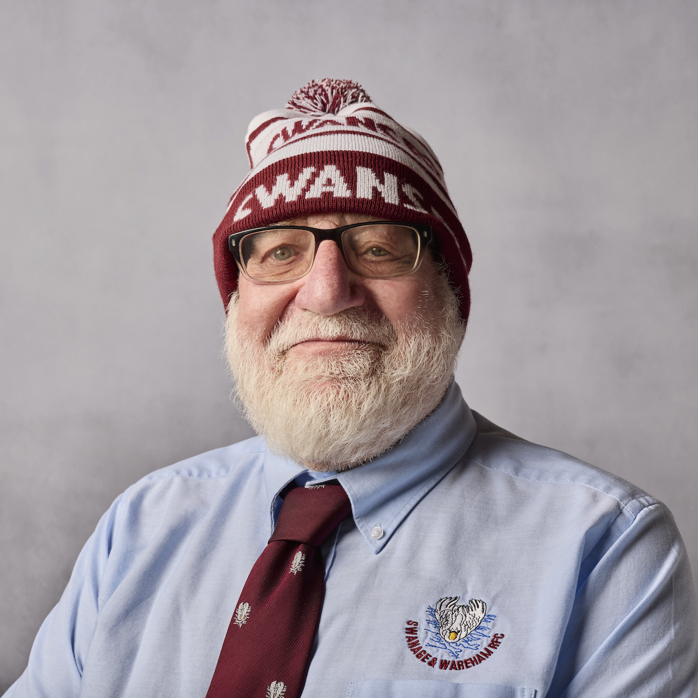 Portrait of an older bearded man wearing glasses, a tie and a knitted bobble hat. The word SWANS is showing at the front of the hat.