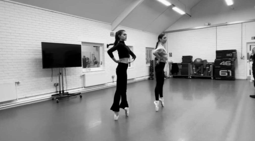 Two dancers in a studio space standing on tiptoe and hands about their hips