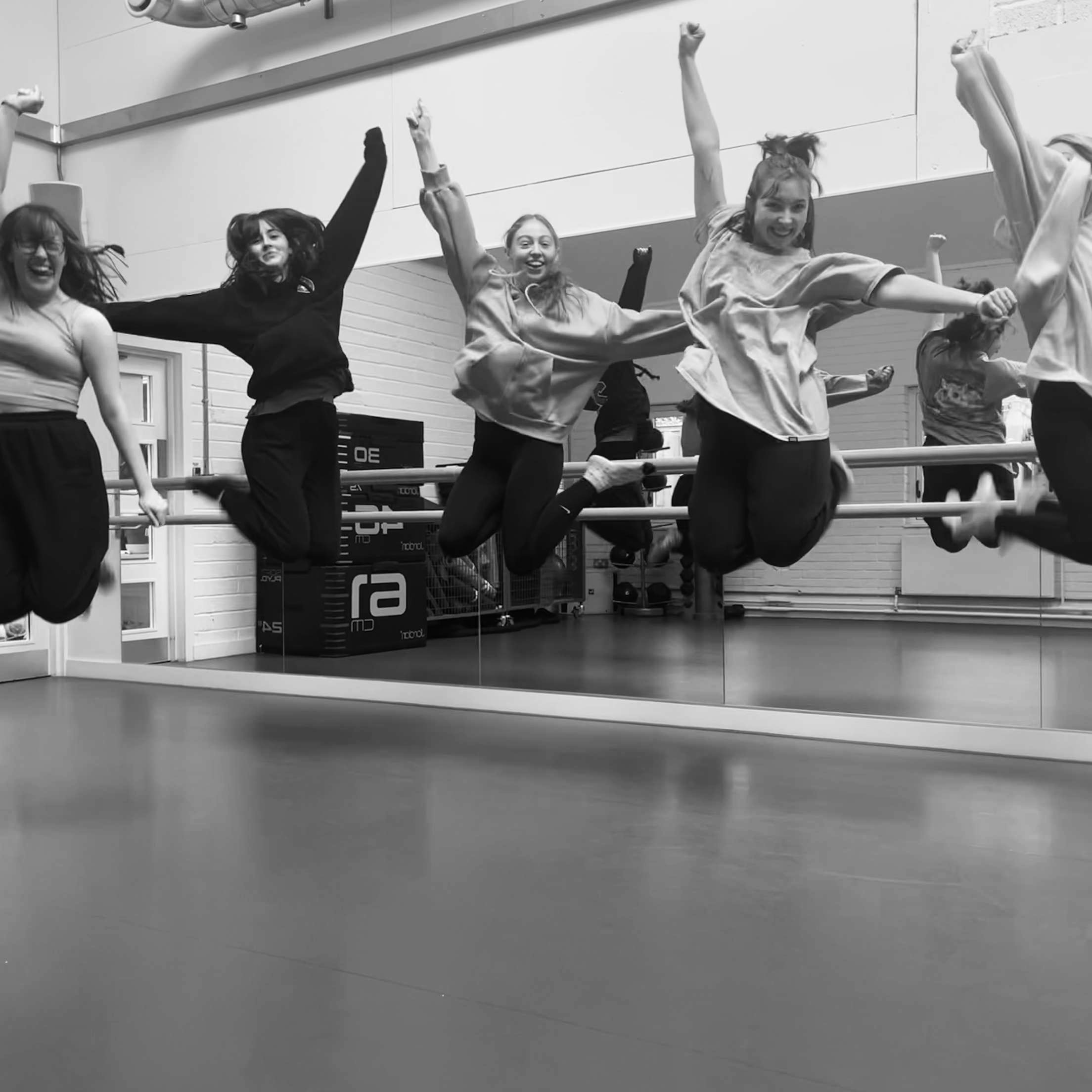 Five dancers in a studio in front of a mirror jump up with their arms outstretched.