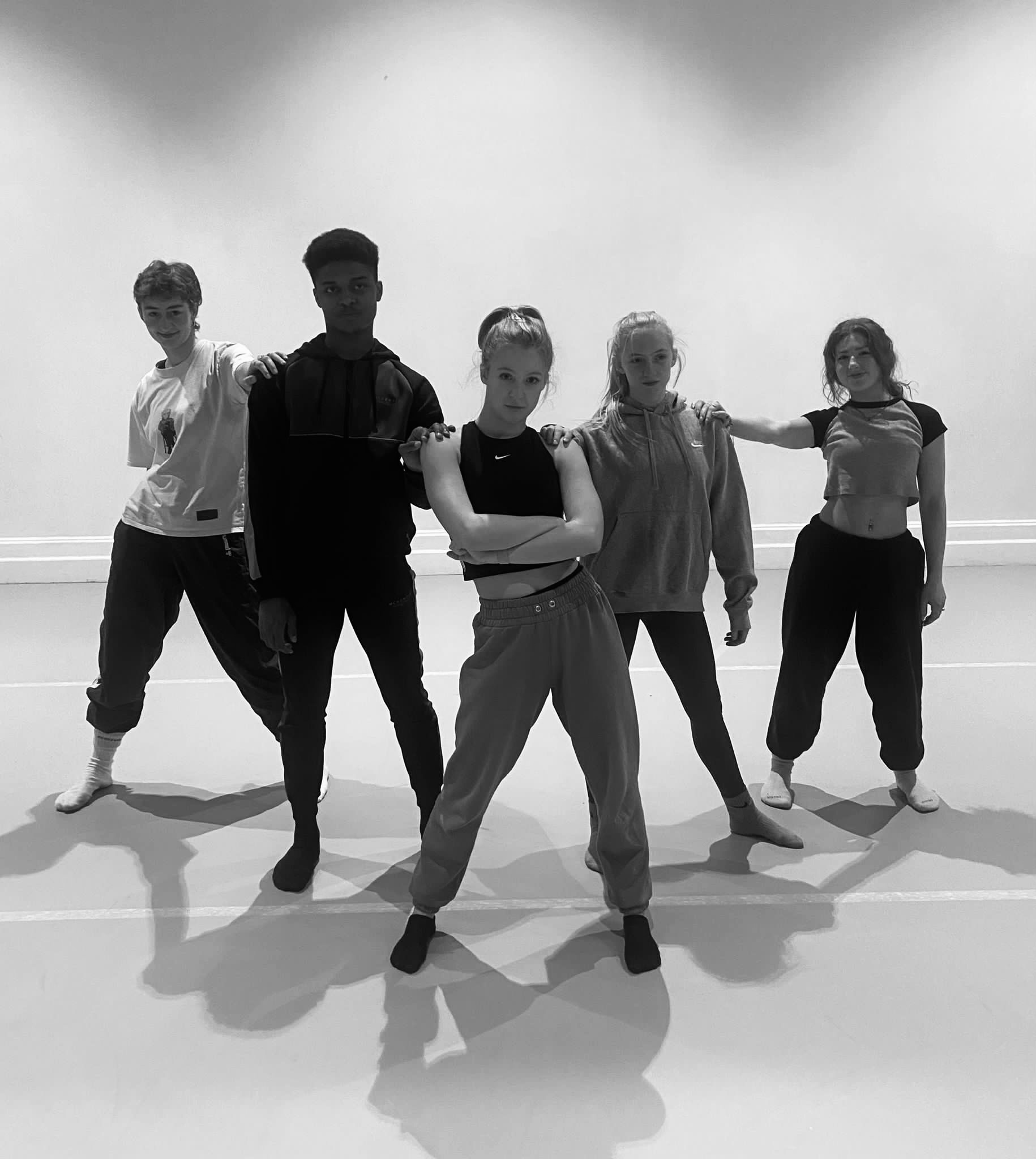 Five dancers stand in a triangular formation in a dance studio and pose for a photo.