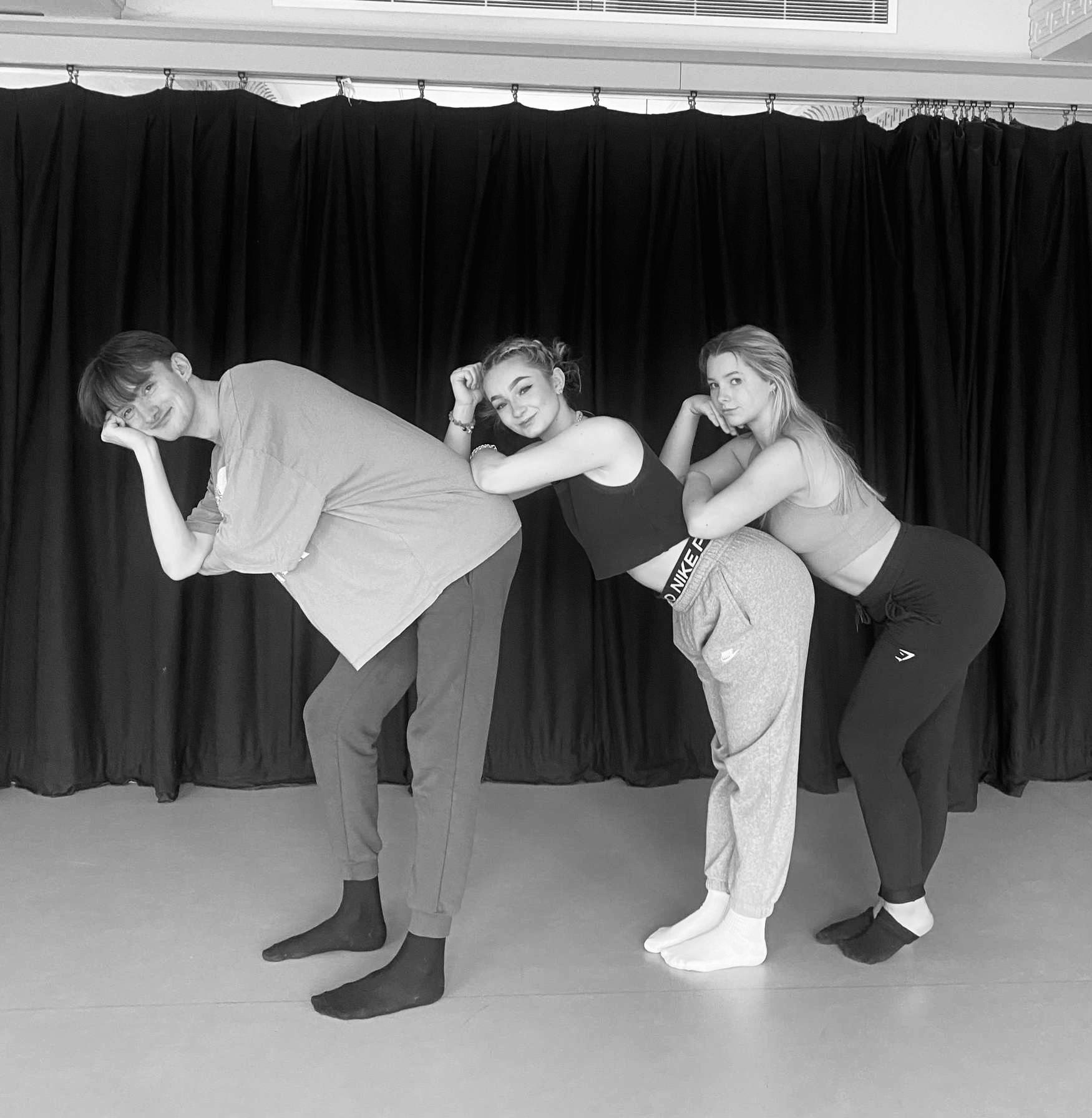 Three dancers lean over on one another in front of a long curtain, with their heads in one of their hands and look towards the camera.