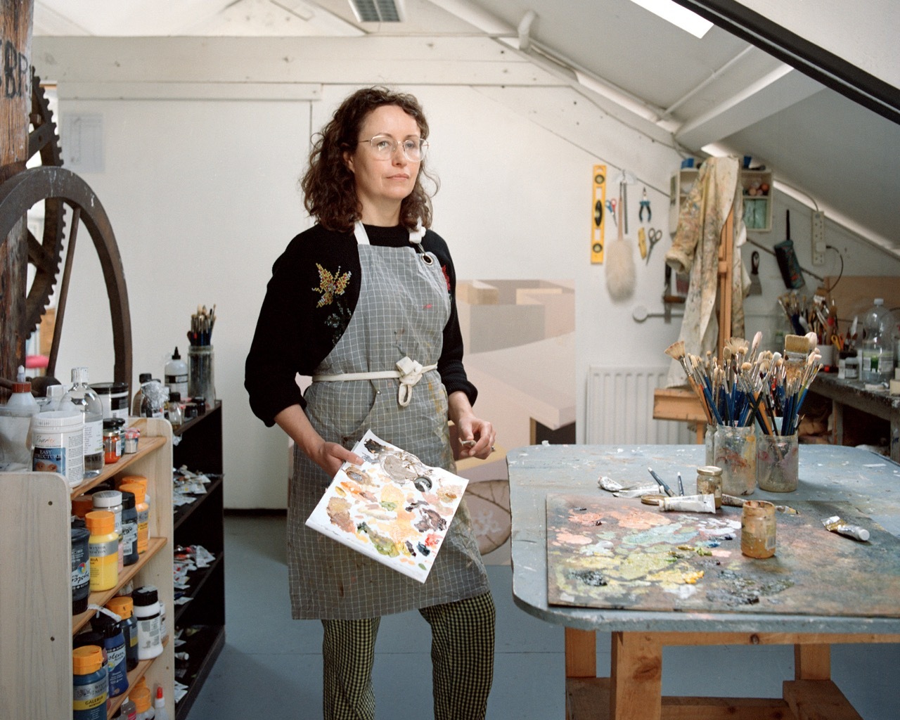Artist Deirde Frost stood in a studio with a grey apron and black top, holding a board of paints next to a paint table with supplies and equipment in the background.