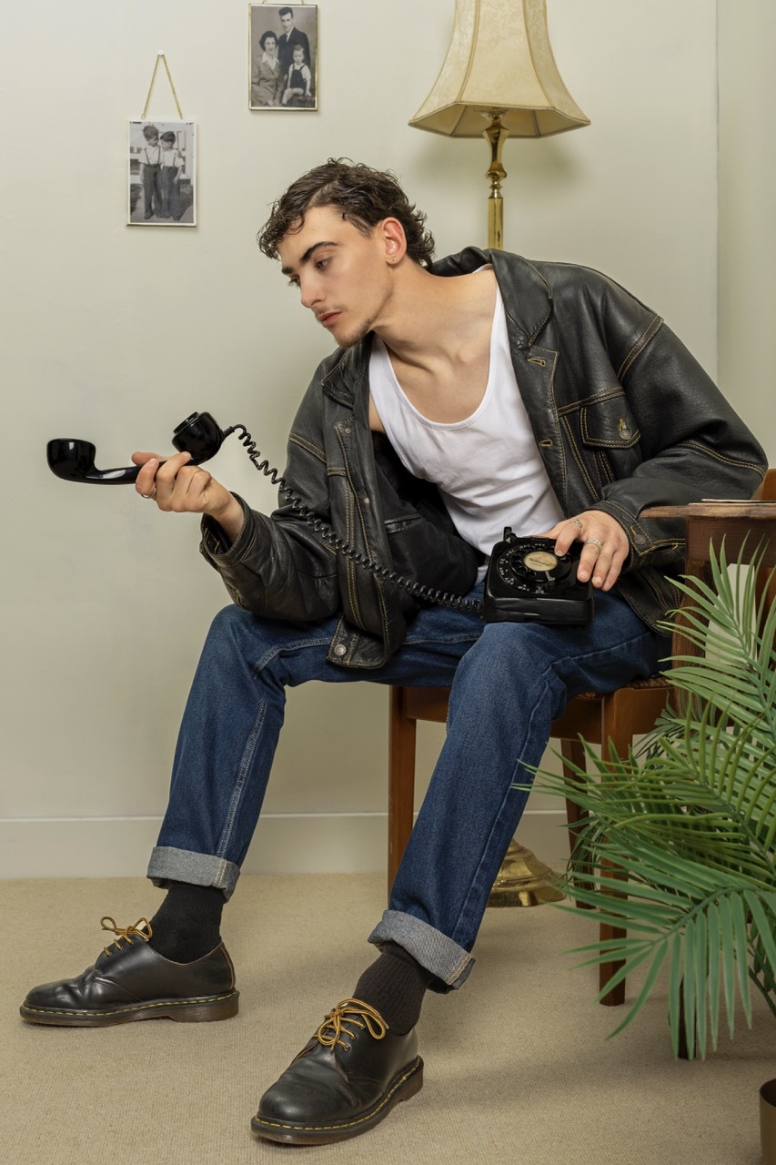 Model in leather jacket and jeans sits on chair holding black rotary telephone, vintage interior with lamp and framed photos on wall.