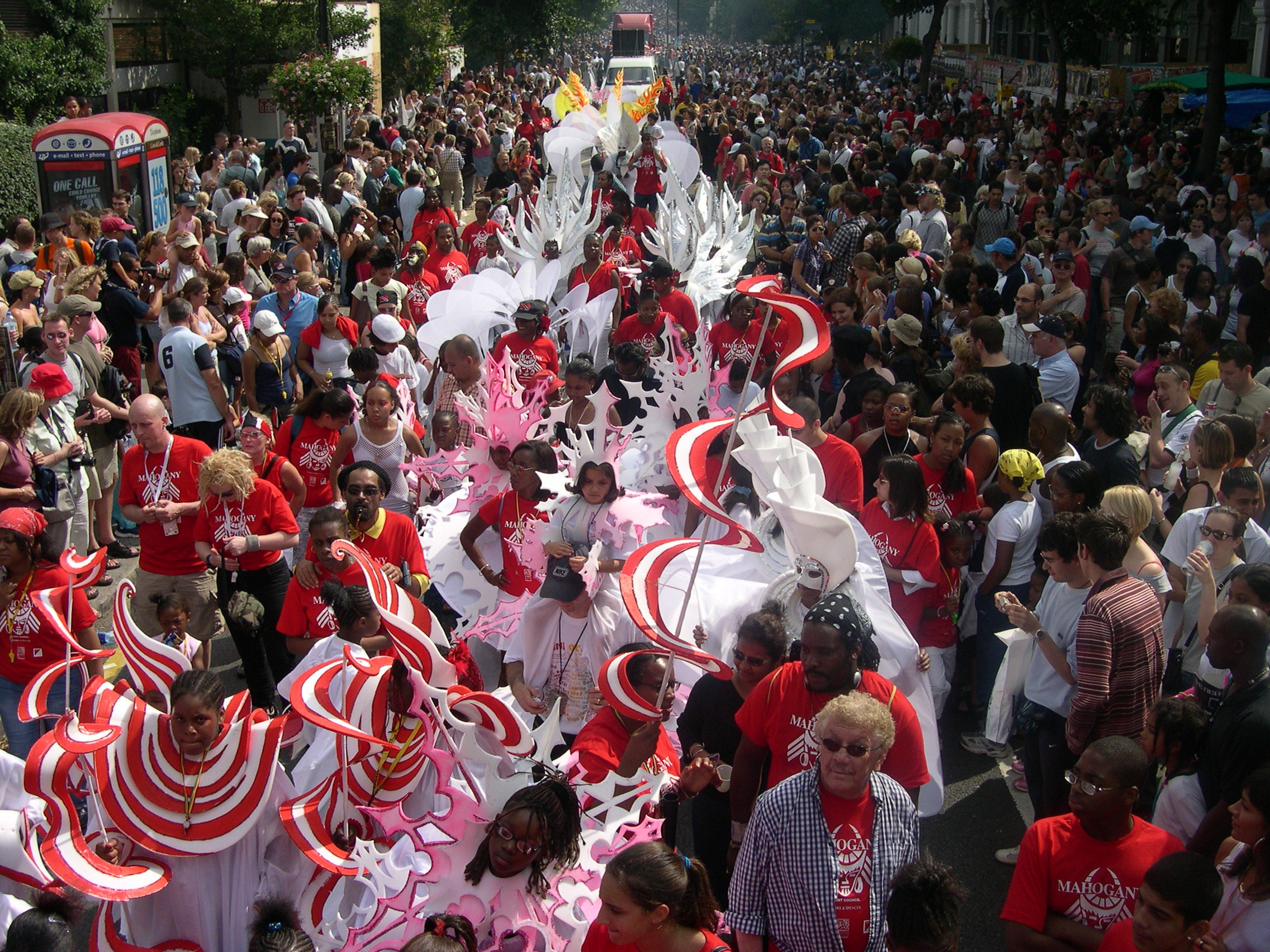A carnival procession with a crowd of performers in red, white and pink costumes, with crowds of spectators either side.