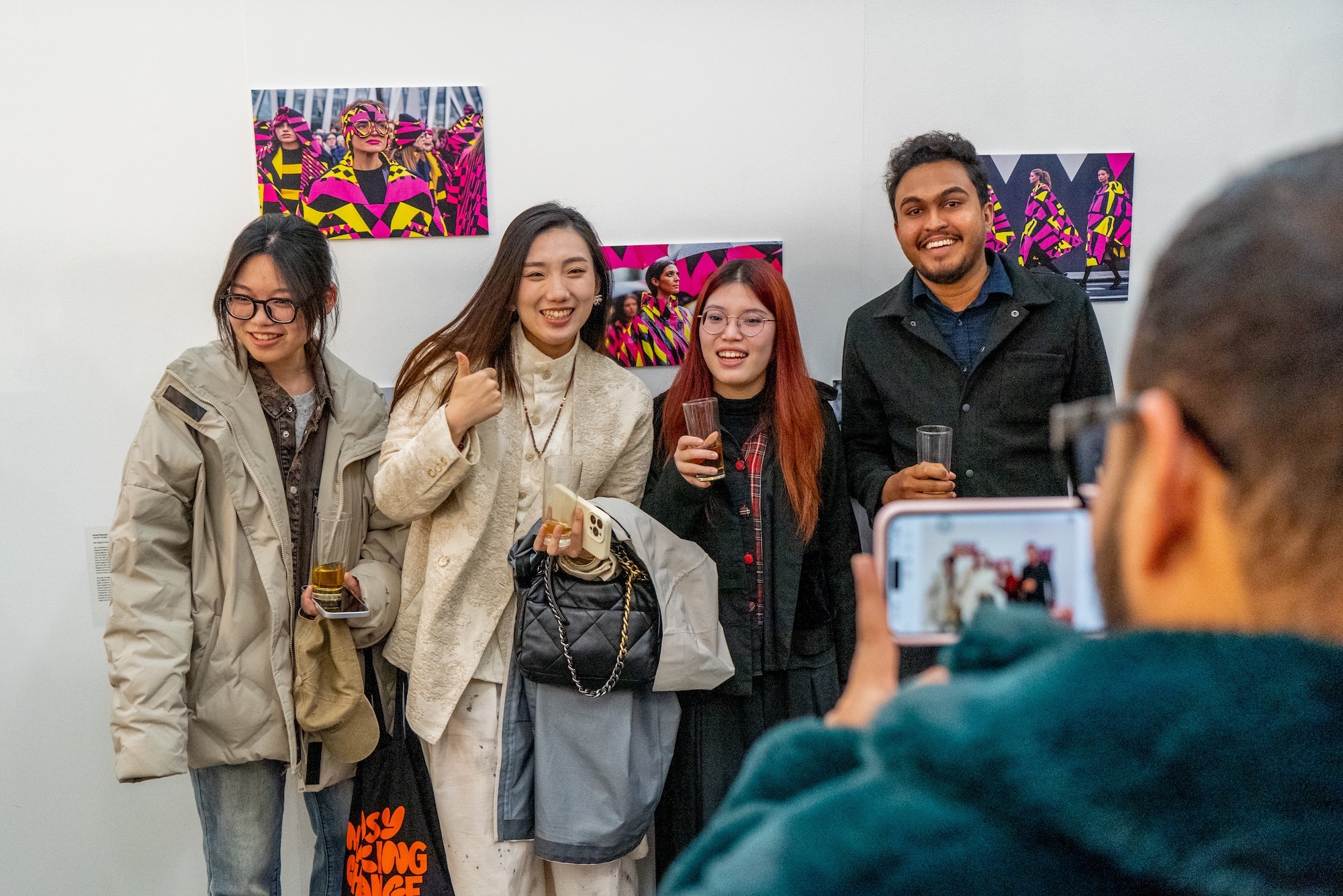 Four people posing together at art gallery opening, colourful artwork on white wall behind them, person taking photo in foreground.