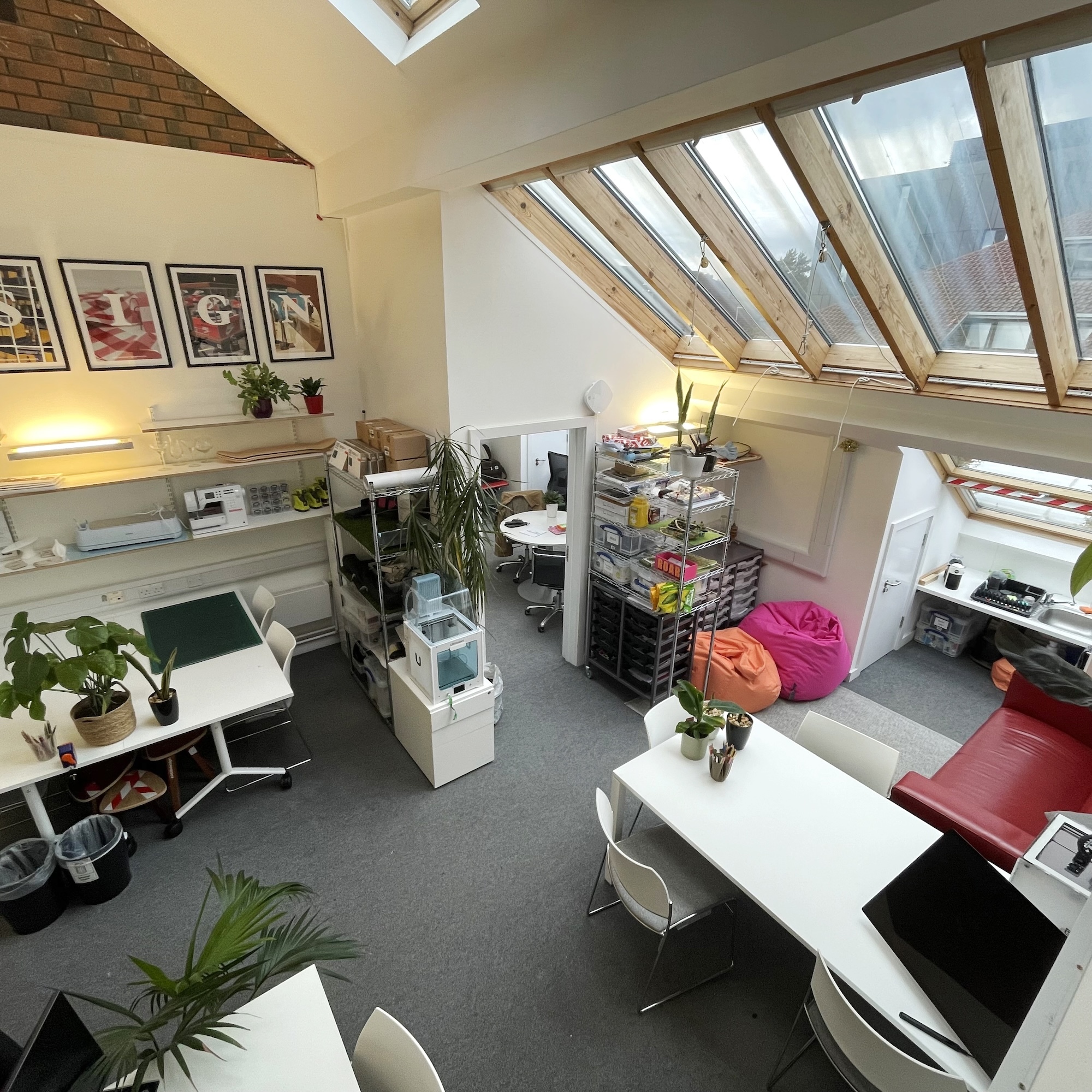 An office space with two desks, each with two chairs by it. There are office shelves and a red sofa also in the room. A large slanting sky light is in the background.