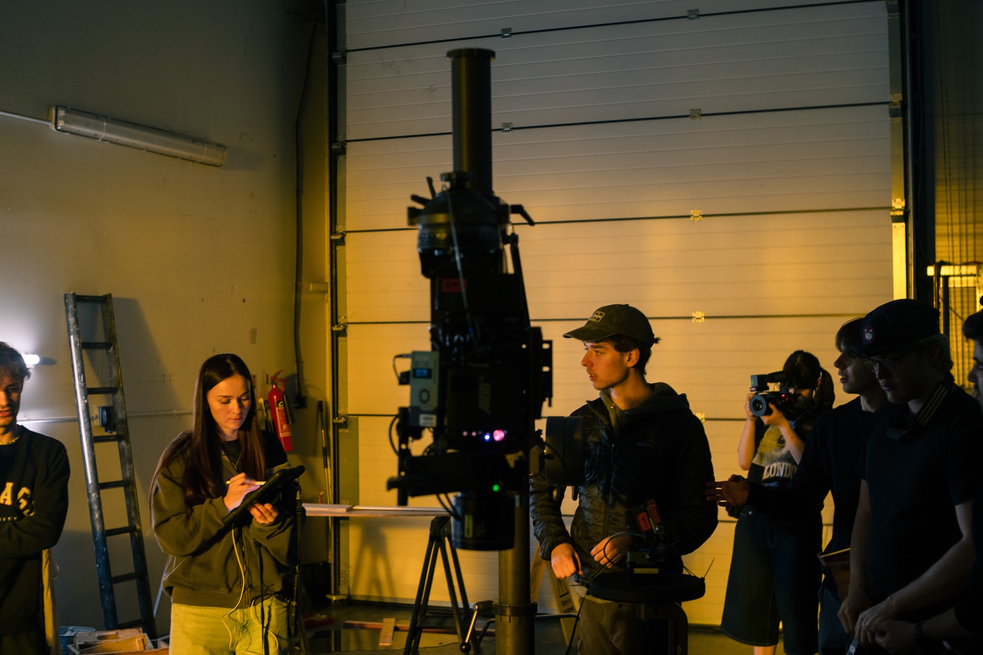 A film crew is gathered in a dimly lit space, with a camera on a tripod at the forefront. A woman with long dark hair is taking notes, while three men look on, one holding a camera. The background features a large garage door and a ladder.
