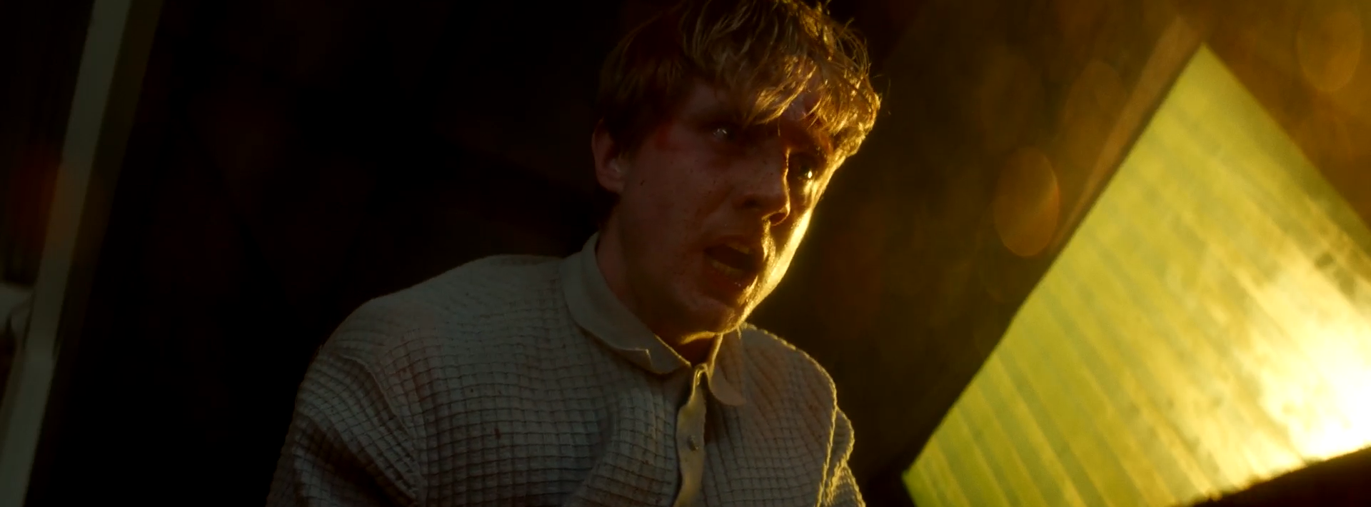 A man with tousled hair wears a light-coloured shirt while sitting in a dimly lit space. A green-tinted window is visible in the background, casting light into the scene, highlighting his facial features.