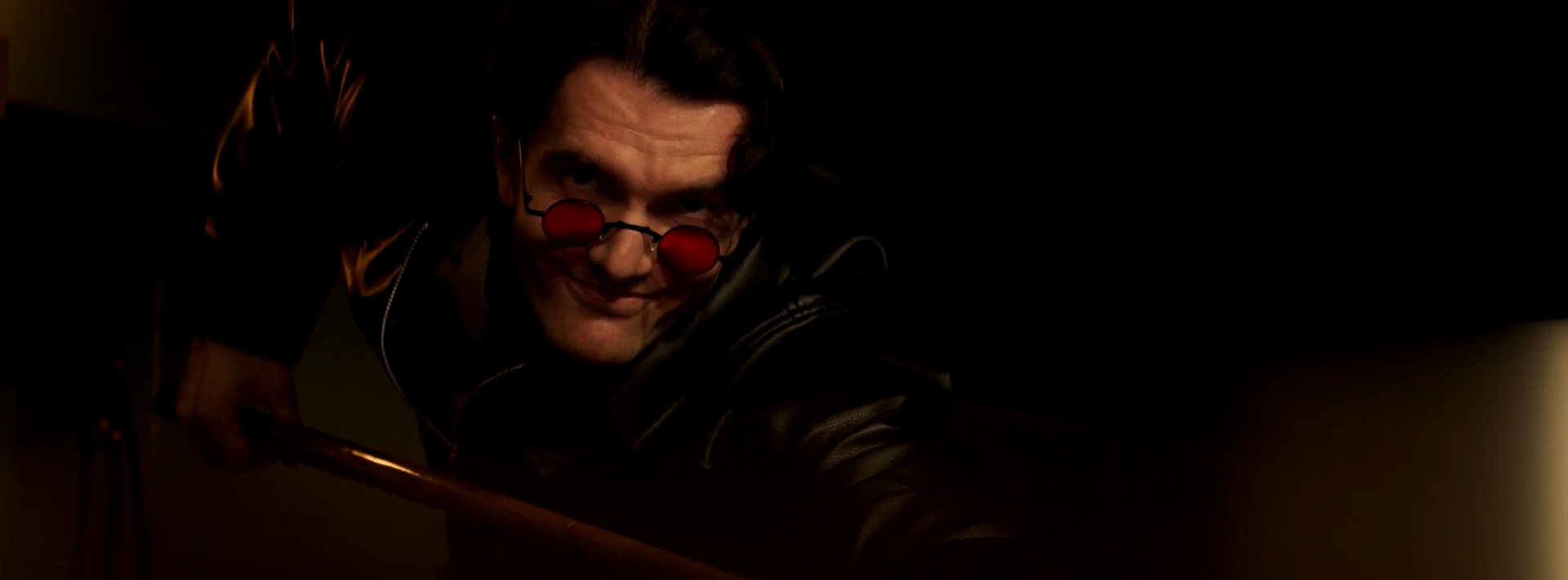 A man with dark hair and red sunglasses is leaning over a wooden surface. He wears a black leather jacket, and the background is mostly dark, creating a dramatic contrast with the soft lighting illuminating part of his face and the nearby object.